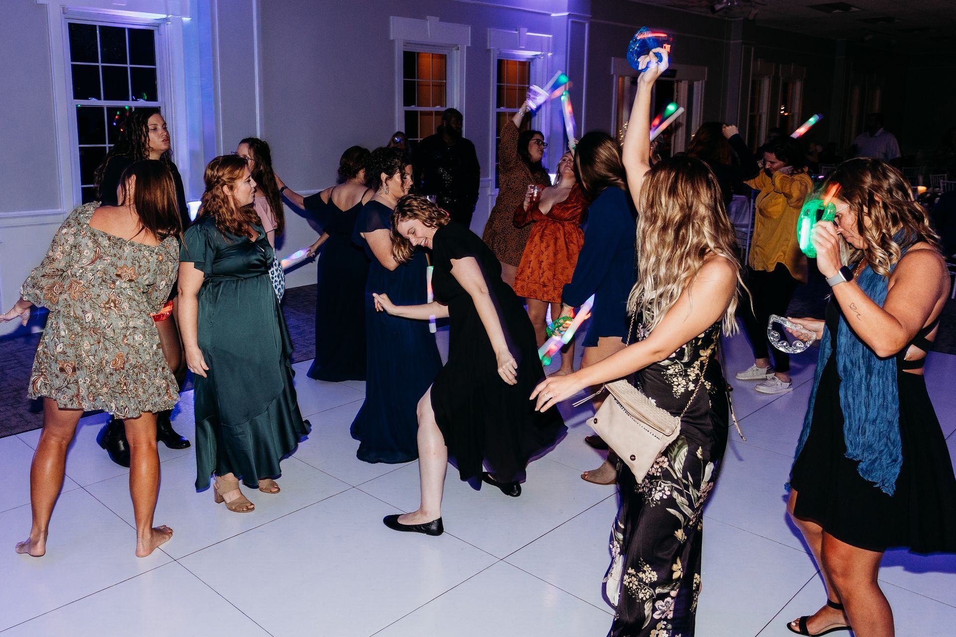 People dancing on a white dance floor, some holding glow sticks, at a party or event, indoor setting.
