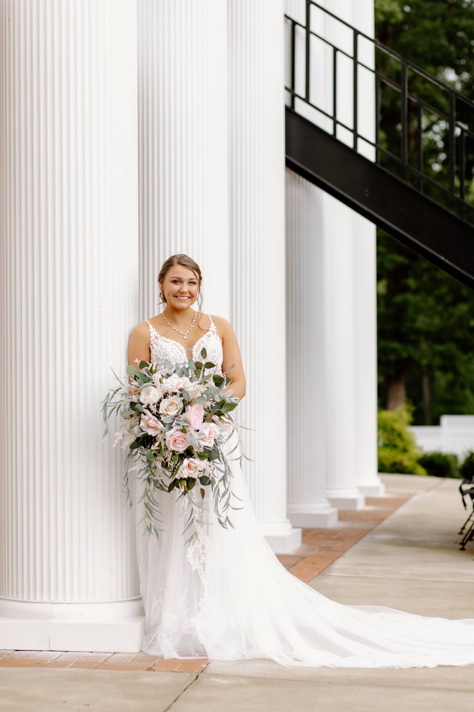 Bride in white wedding dress holding bouquet, smiling by white columns.