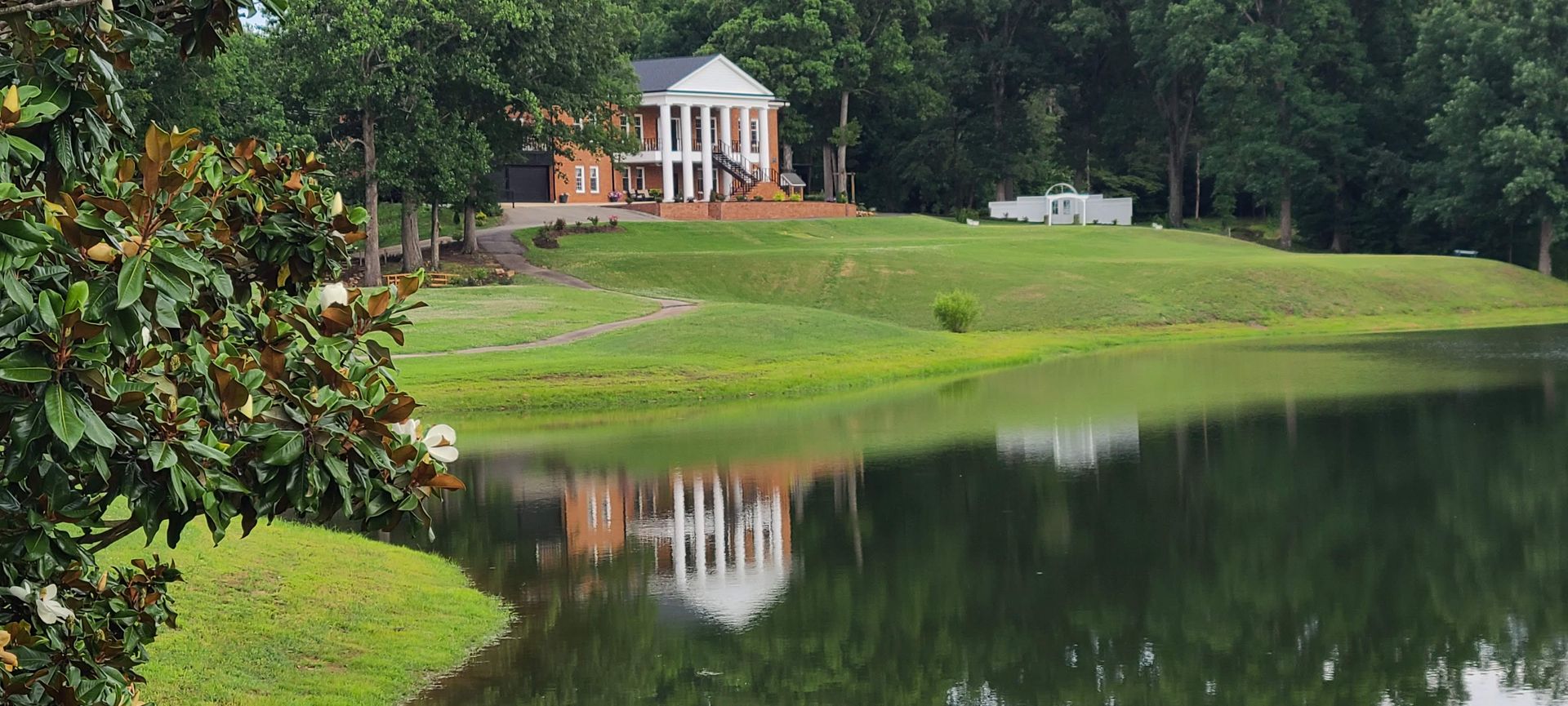 A stately home with white columns reflects in calm water. Green grass and trees frame the scene.