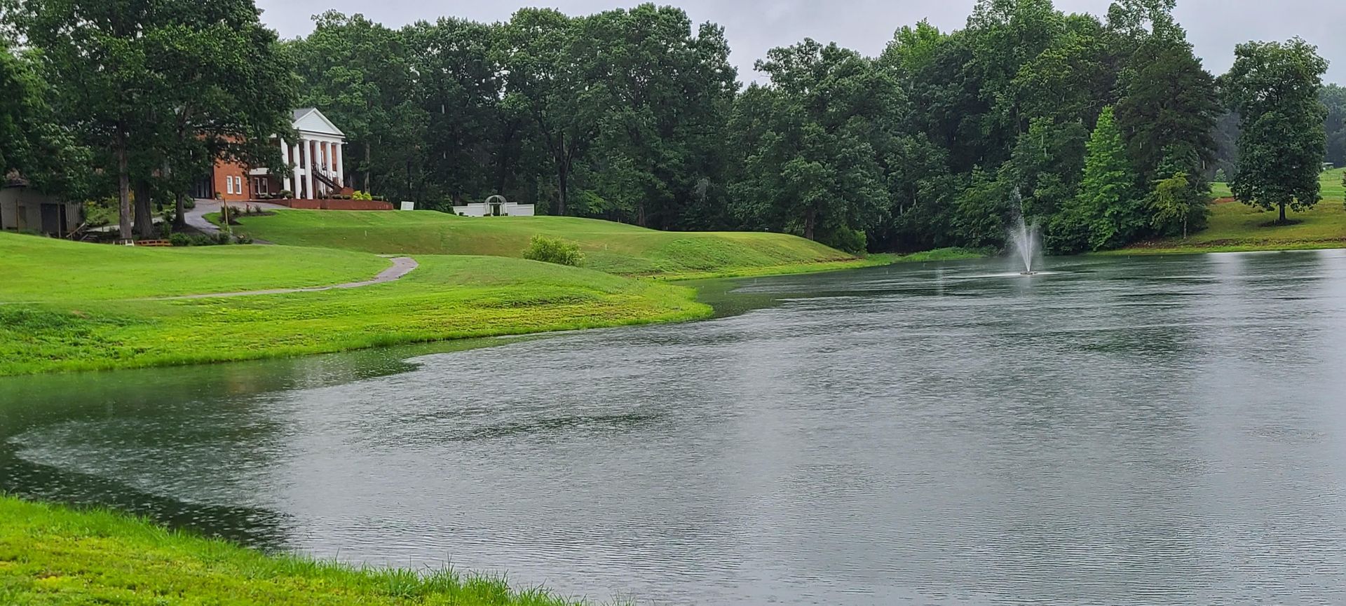 A rainy day scene. A body of water with a fountain in the distance and a house near green grass and trees.