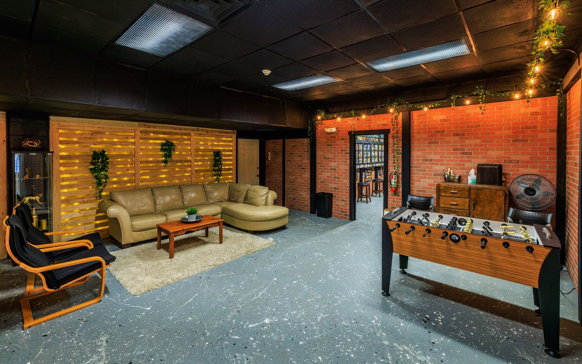 Lounge area with sofa, foosball table, and exposed brick walls. Black ceiling with lights and decorative plants.