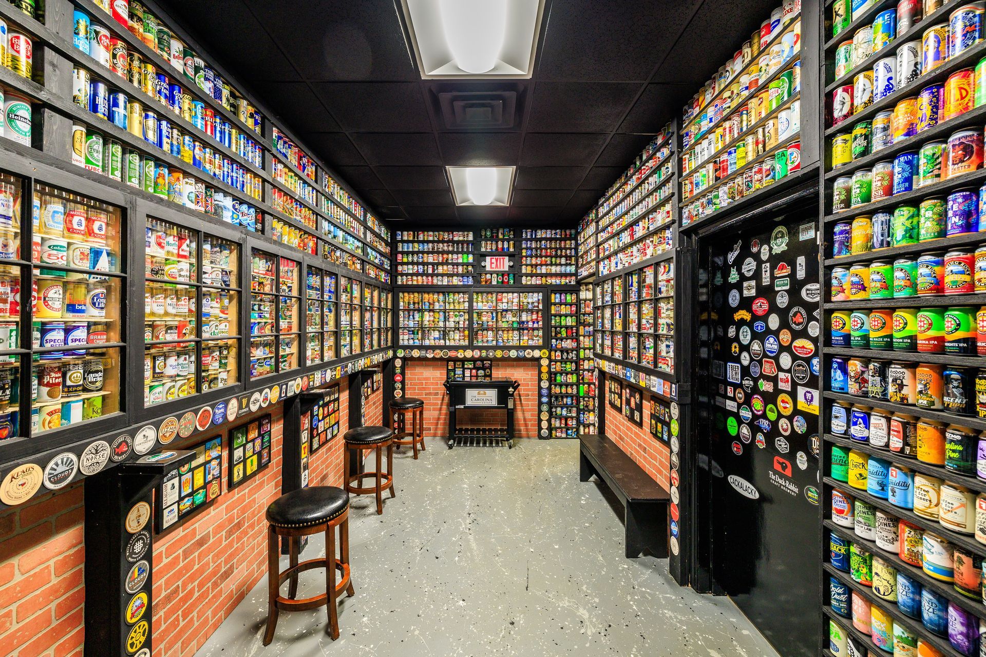 Narrow room lined with beer cans on shelves, bar stools, brick accents, black ceiling and door.