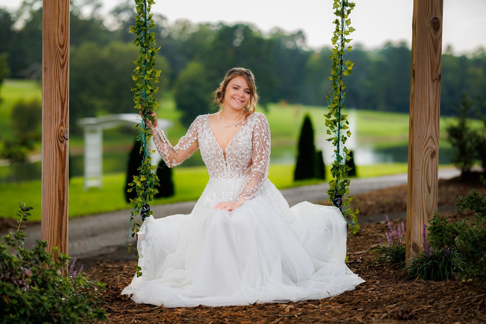 Bride in a white dress sits on a swing, smiling. Background includes a pond and trees.