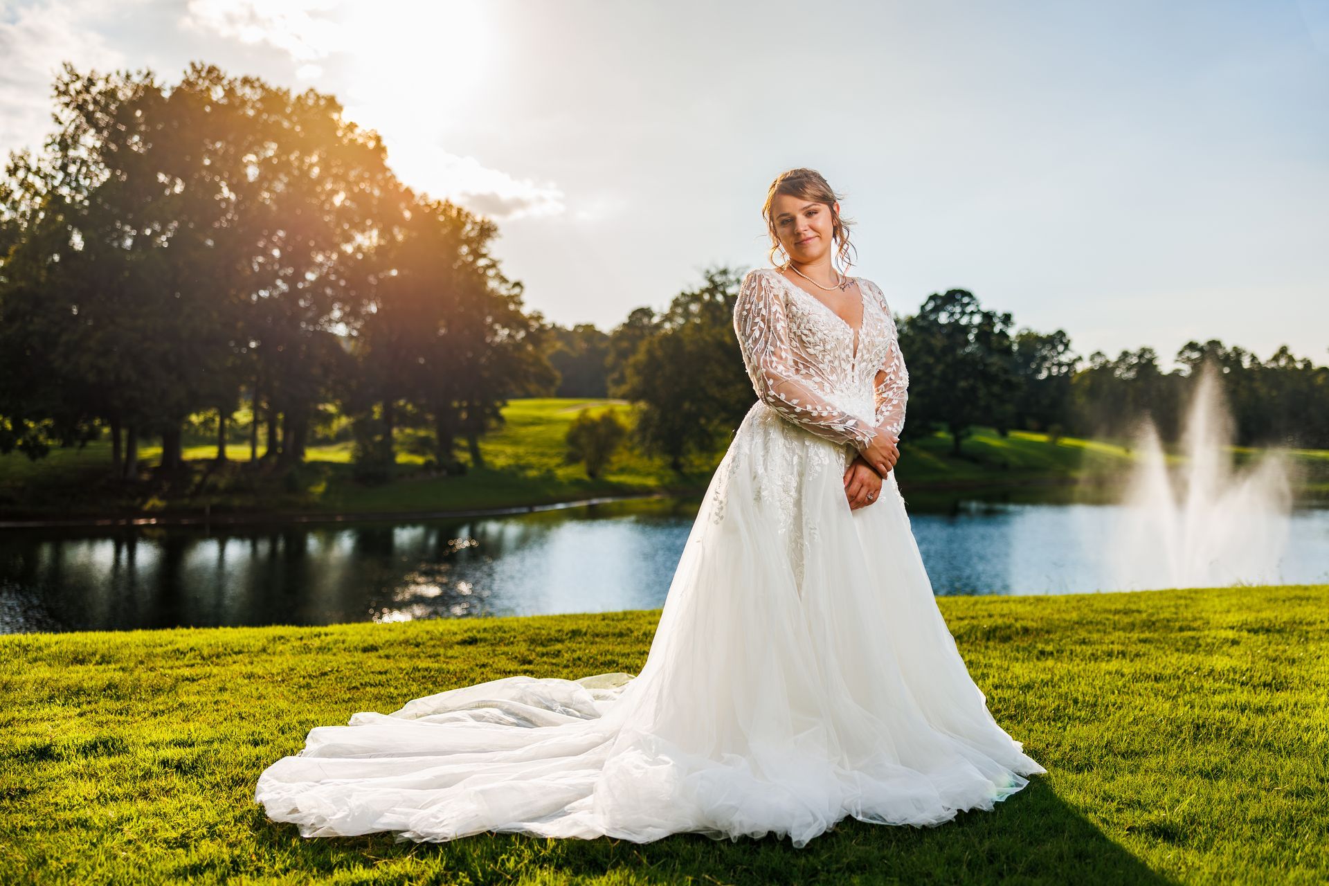 Bride in white dress on green grass, posing near a lake with fountains, sunny day.