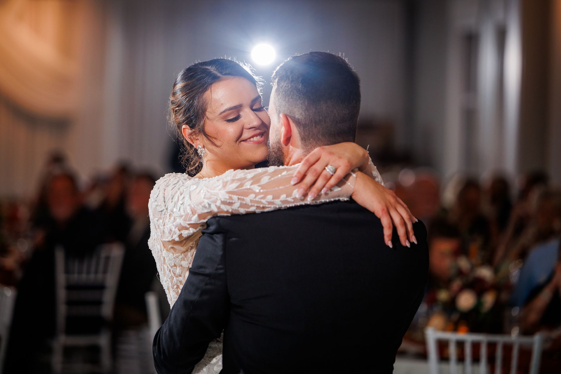 Bride and groom embrace on a dance floor, smiling, with a bright light overhead.