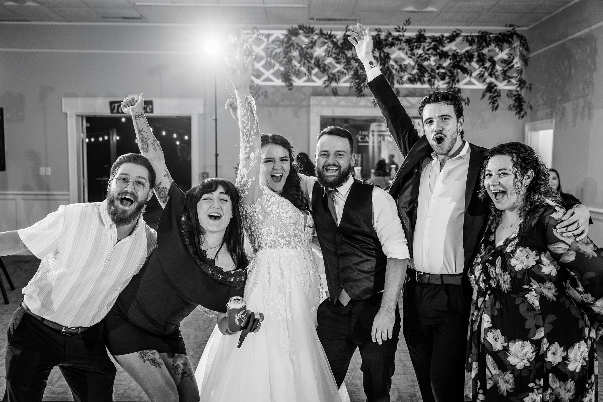 Wedding party celebrating indoors with arms raised, joyous expressions, and a bright light.