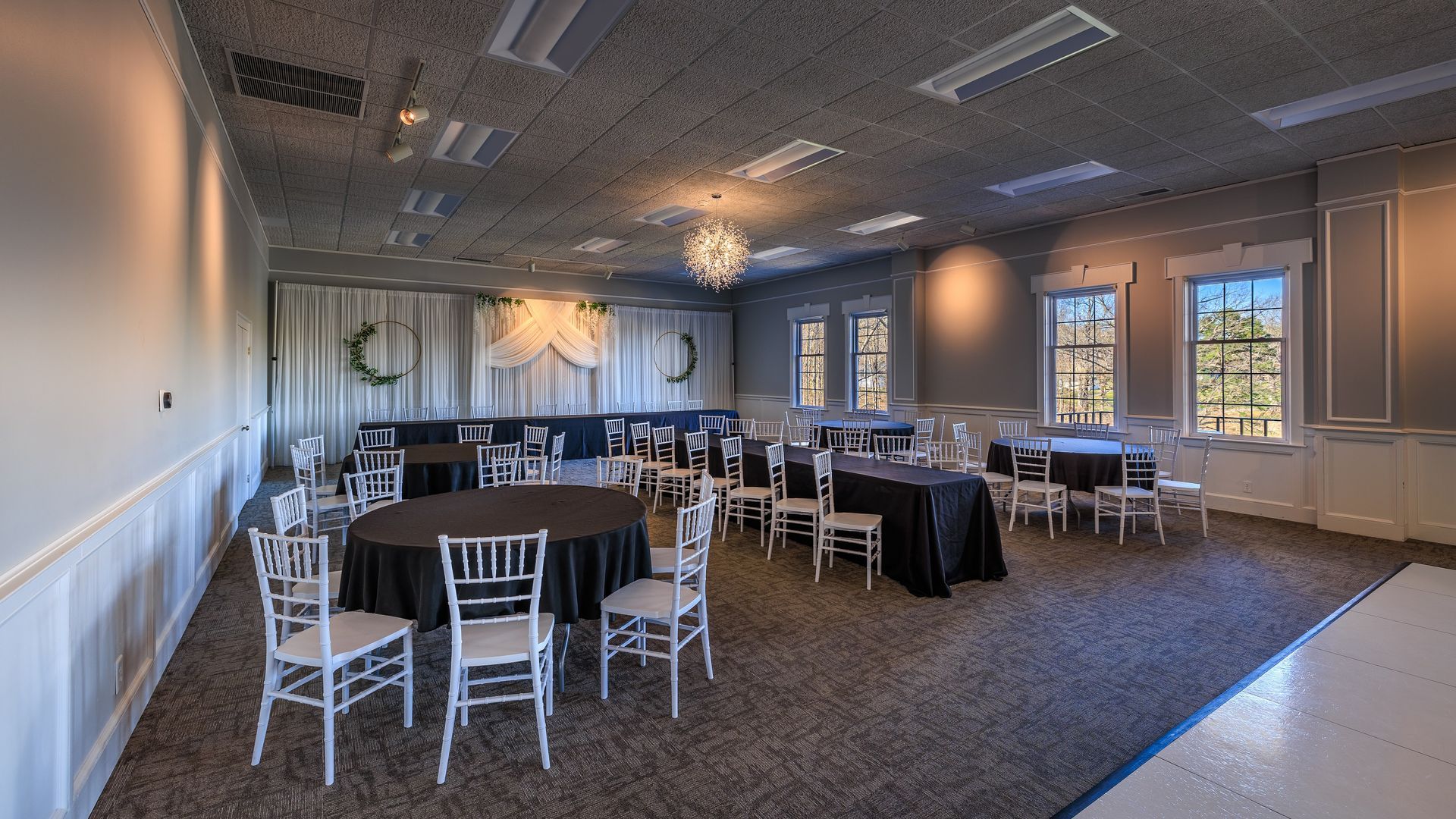 Banquet hall with round tables covered in black linens, white chairs, and a chandelier.