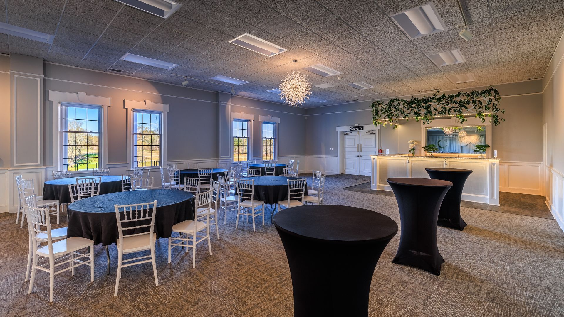 Event space with round black tables, white chairs, and a bar area with greenery.