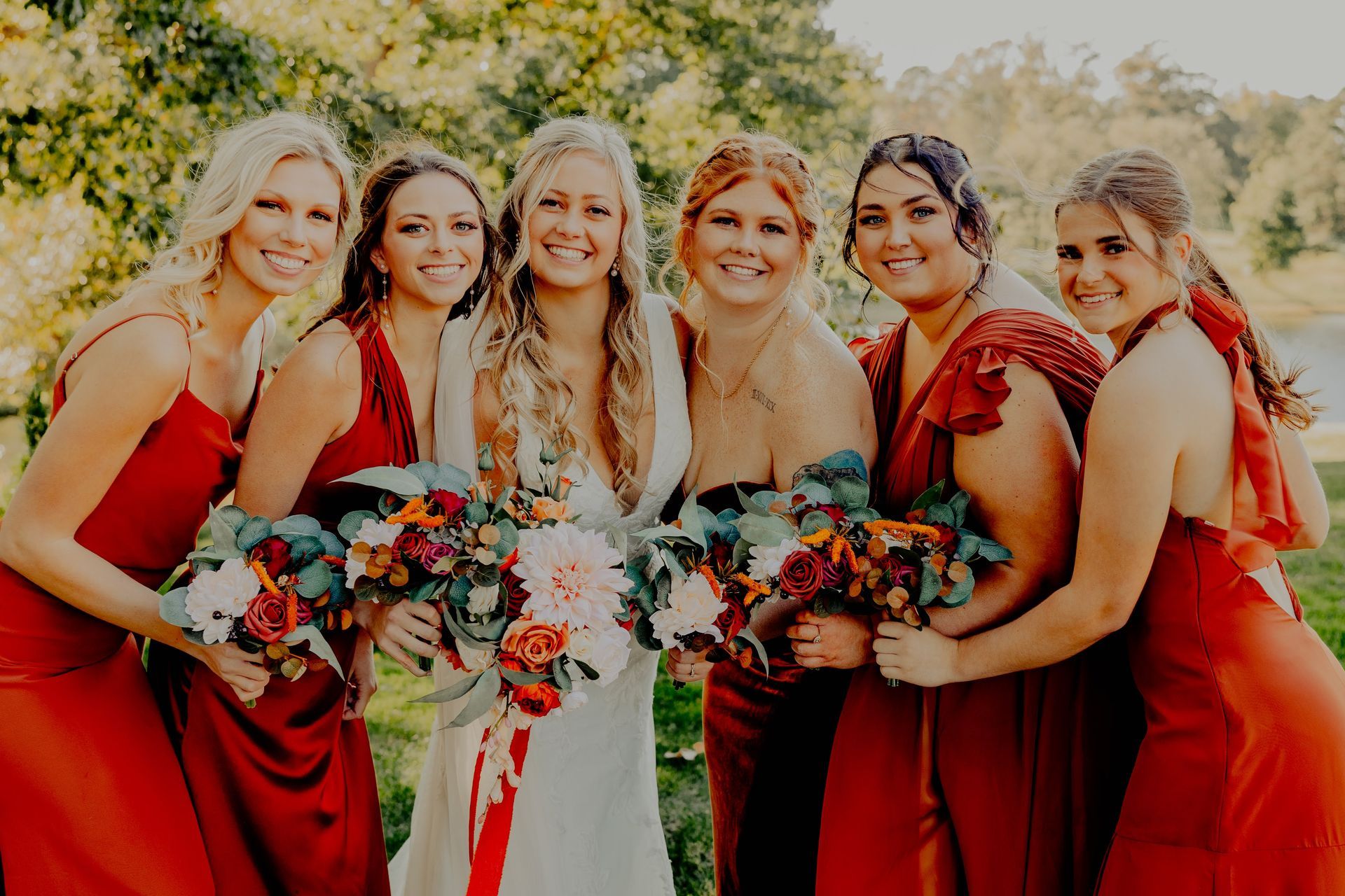 Bridesmaids and bride pose outdoors, holding bouquets. They wear red dresses, smiling.