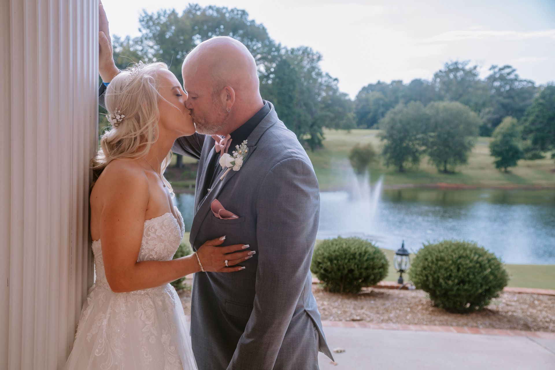 Couple kissing, bride in white gown, groom in grey suit, by a lake with a fountain and trees.