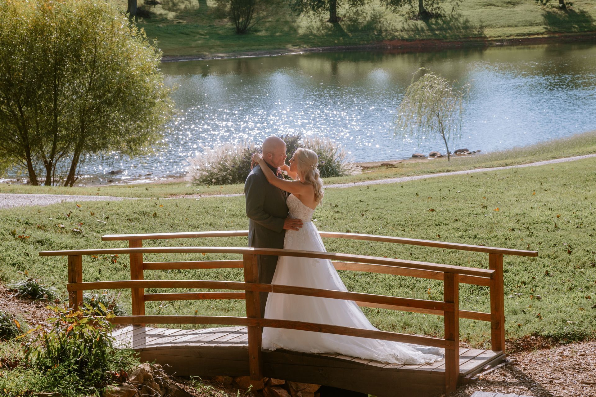 Couple kissing on a wooden bridge overlooking a lake on a sunny day. Bride in white dress, groom in dark suit.