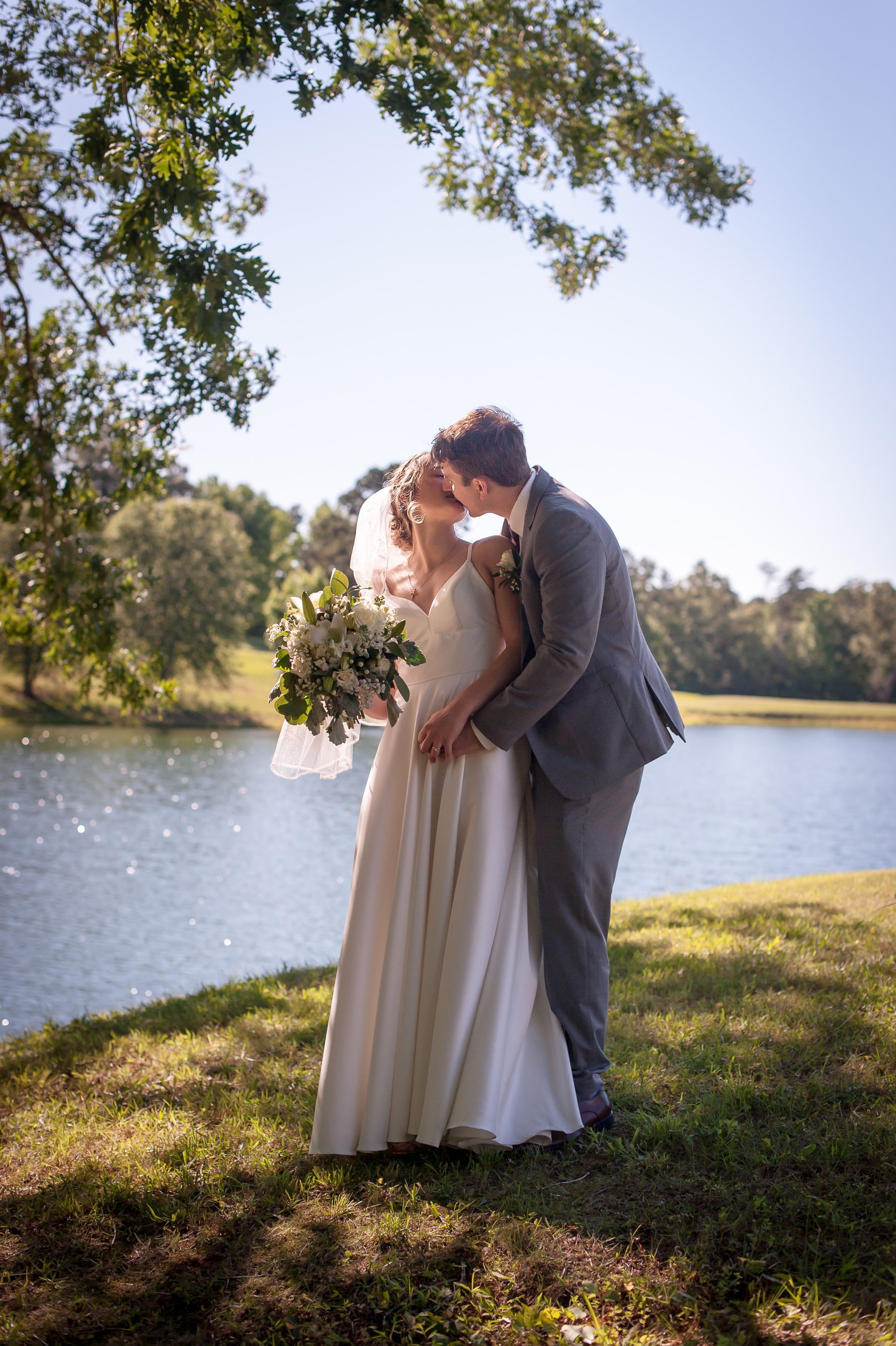 Newlyweds kissing by a lake. Bride in a white gown, groom in a gray suit, under a tree on a sunny day.