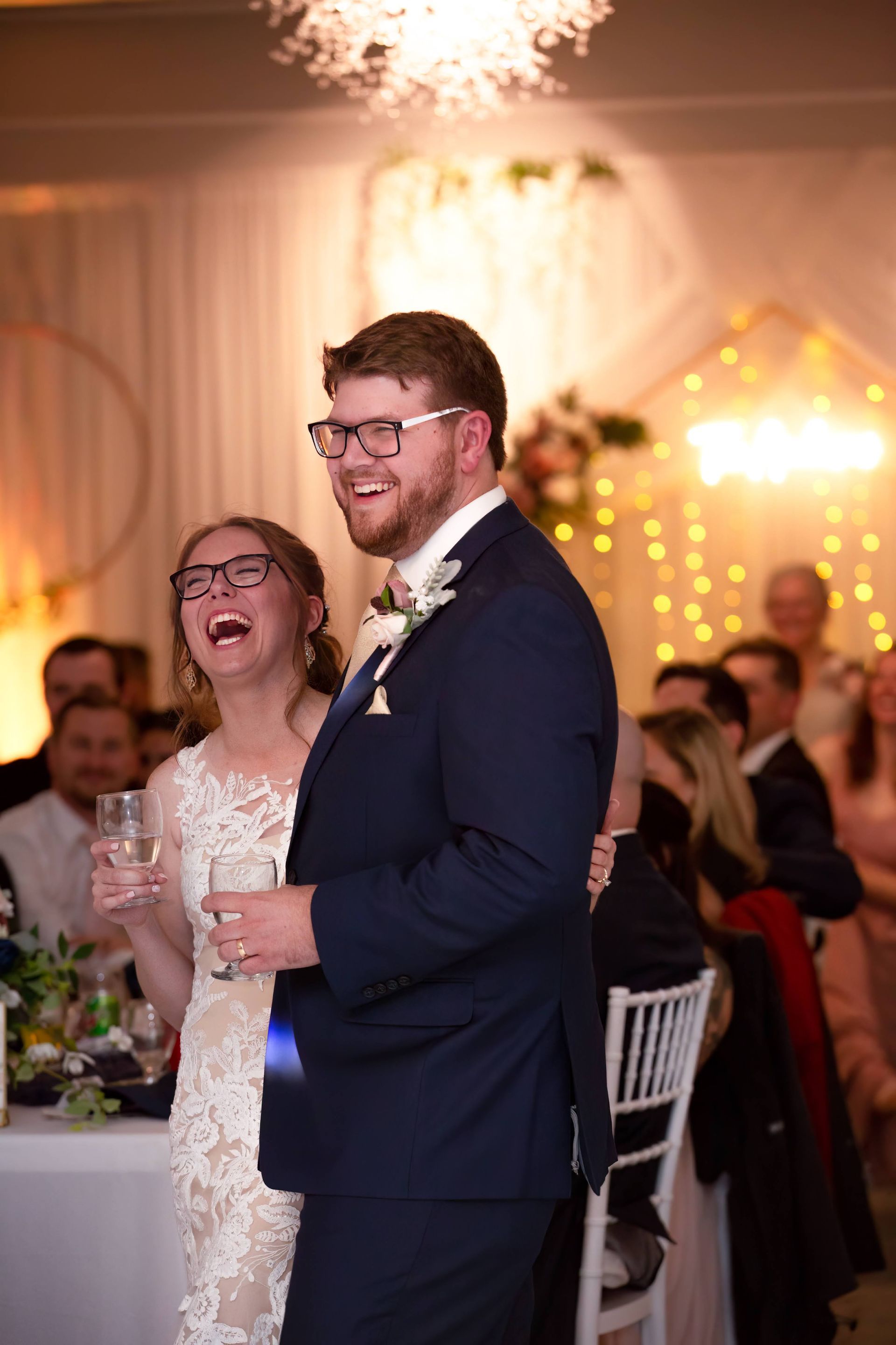 Bride and groom laughing at a wedding reception. She wears a lace dress, he wears a blue suit.