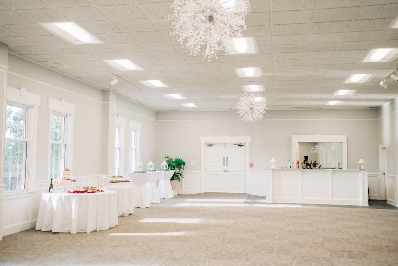 Empty, bright, white-walled event space with a bar, tables, chandeliers, and large windows.