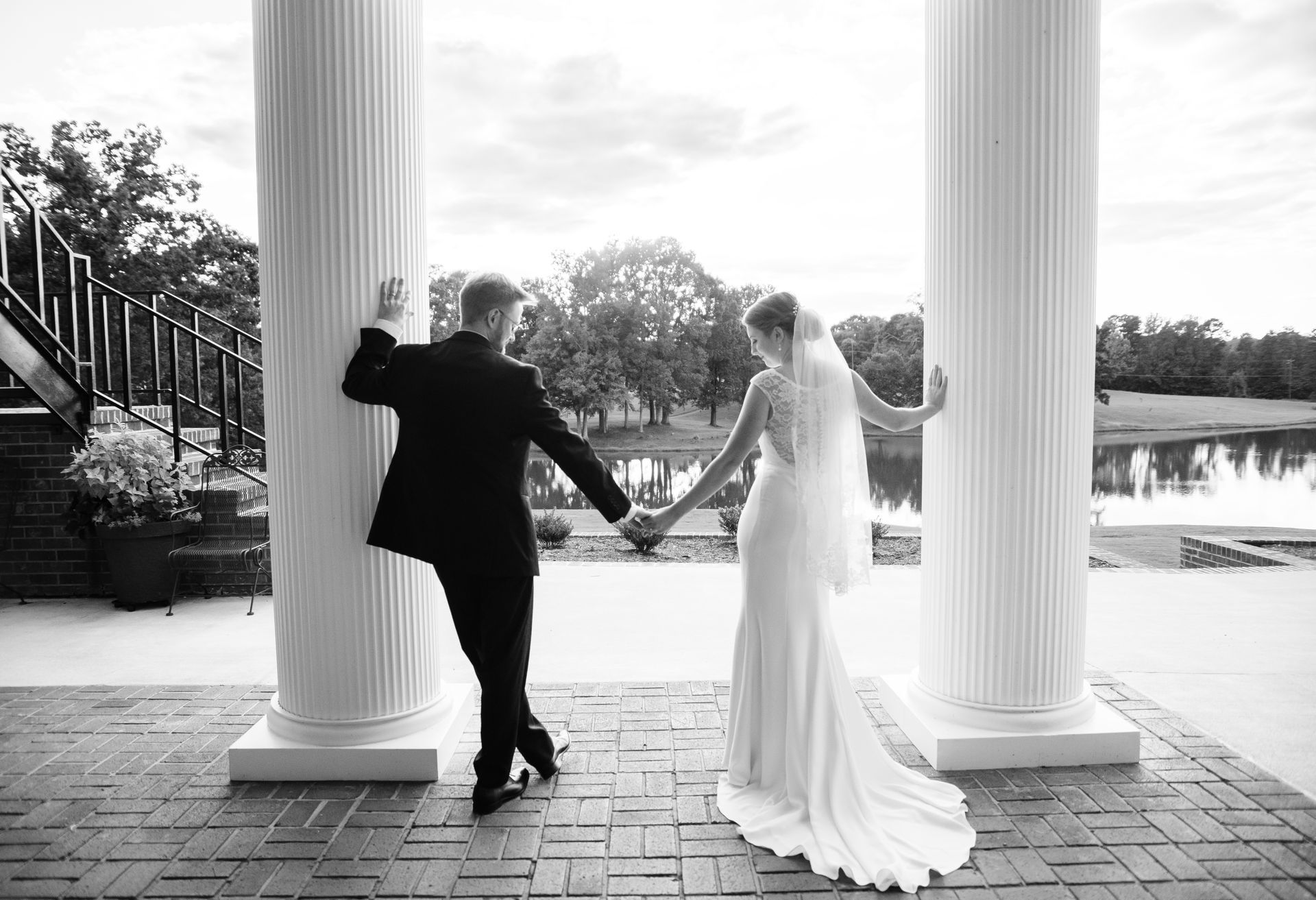 Bride and groom holding hands, leaning against pillars, looking toward lake.