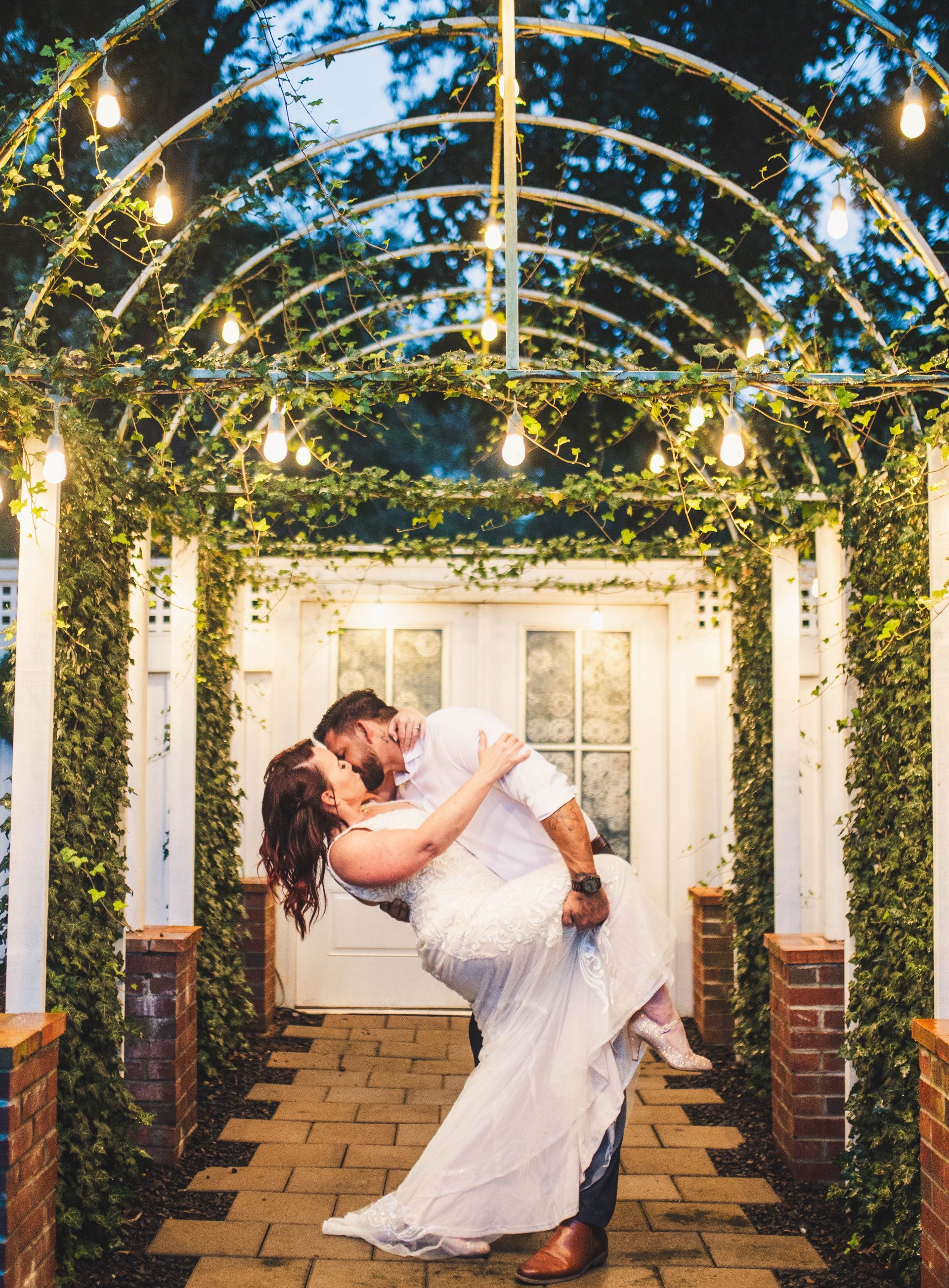 Couple kissing under a string-lit archway, embracing. Ivy and brick accents.