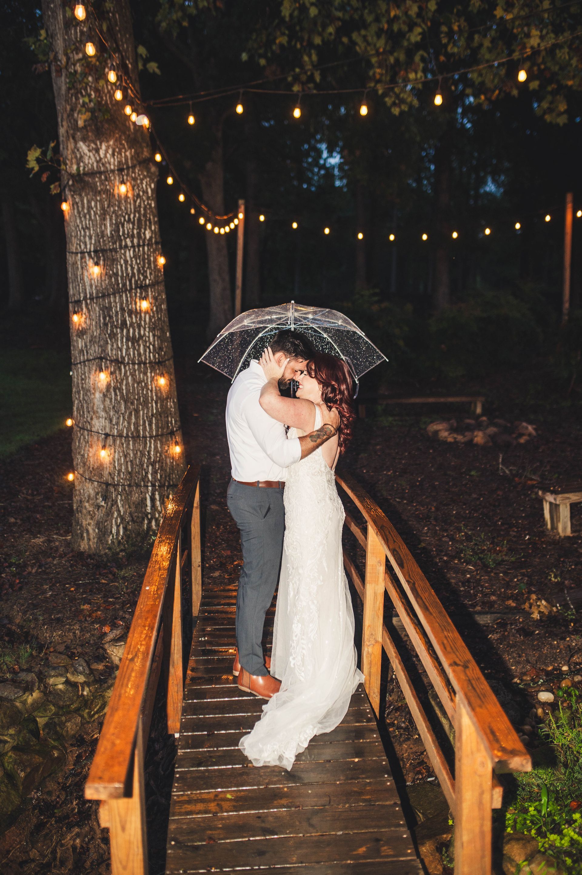Couple kissing under an umbrella on a wooden bridge, strung with lights, in the rain.