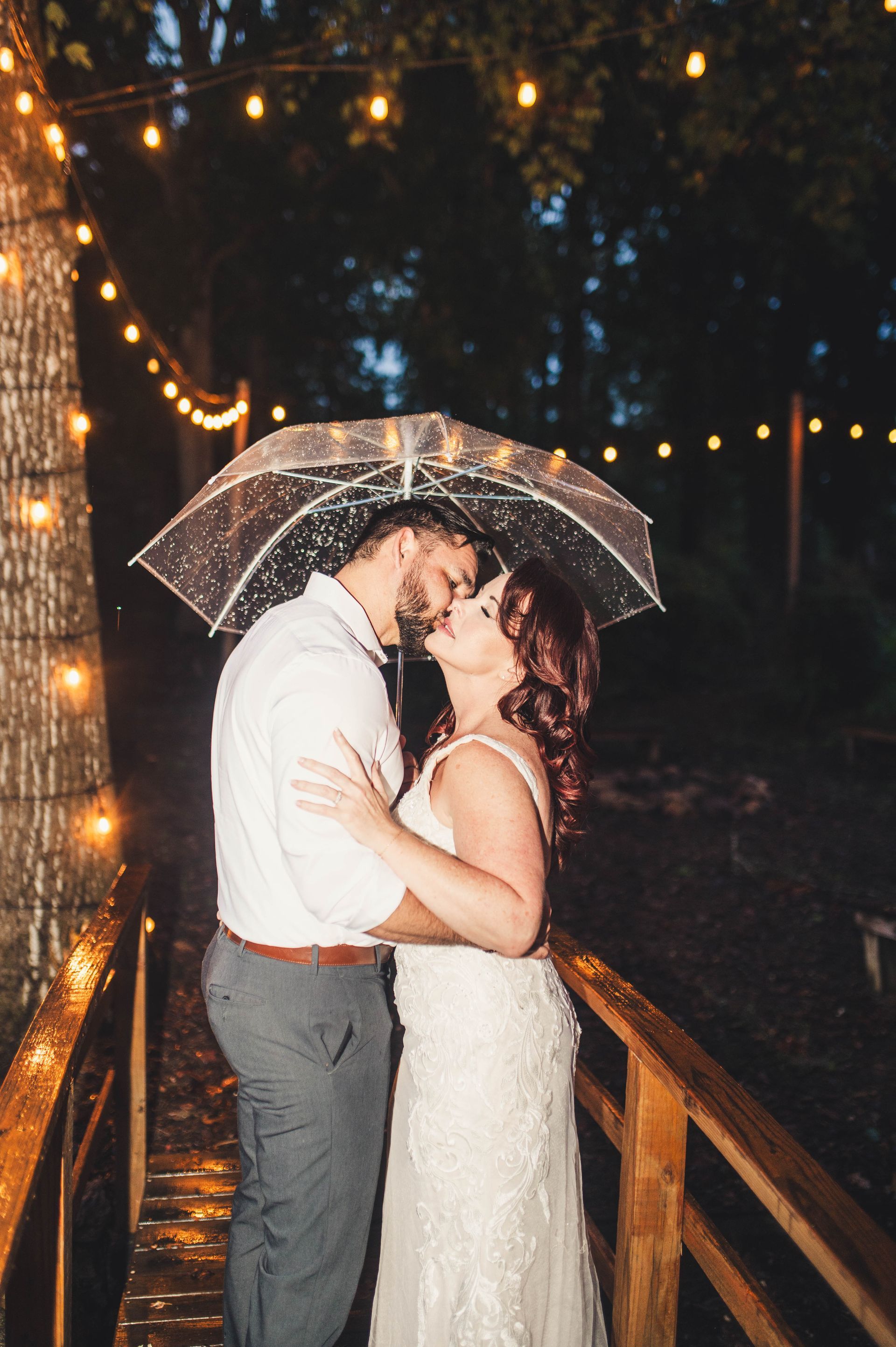 Couple kissing under a clear umbrella on a wooden bridge, rain falling, string lights overhead.