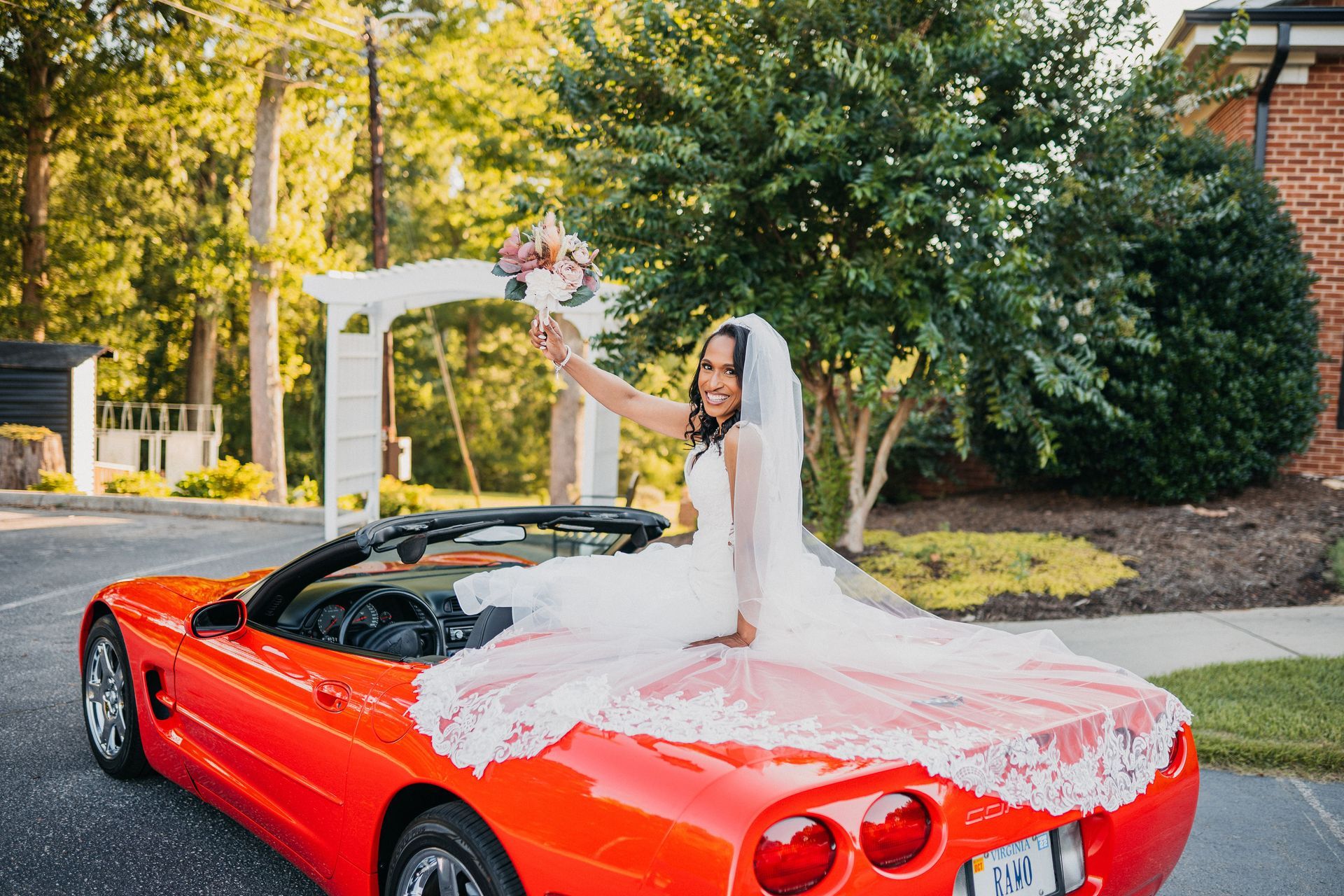 Bride in white dress waves from red convertible, holding bouquet.