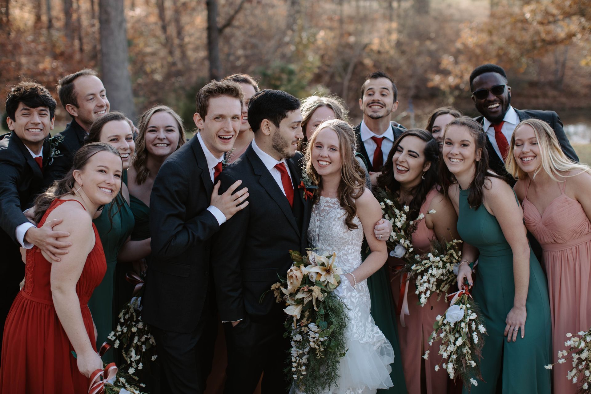 Wedding group portrait: Bride and groom embrace, surrounded by bridesmaids in various colored dresses, groomsmen in suits.