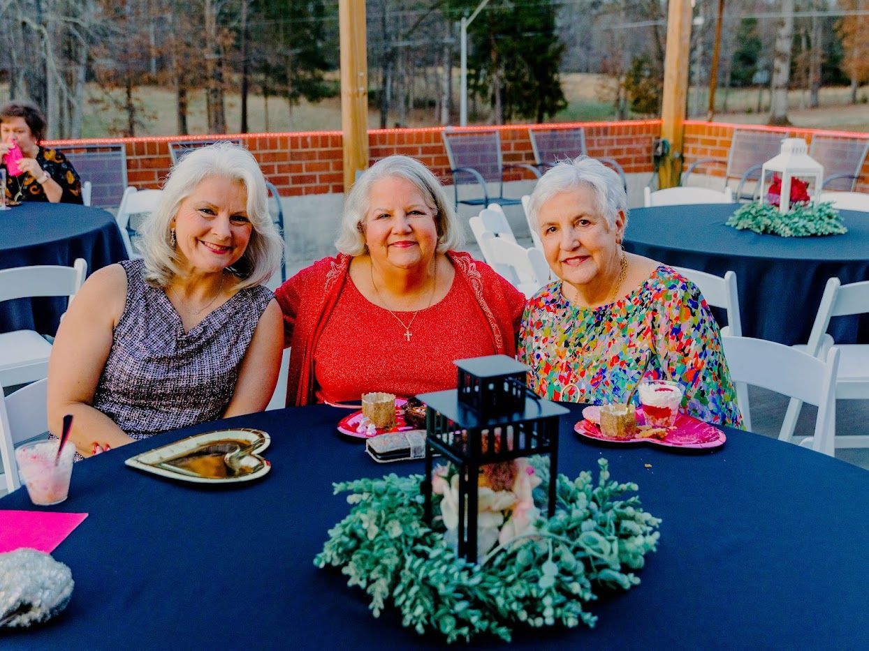 Three women at a table, smiling. Outdoor setting with blue tablecloths, desserts, and decorations.