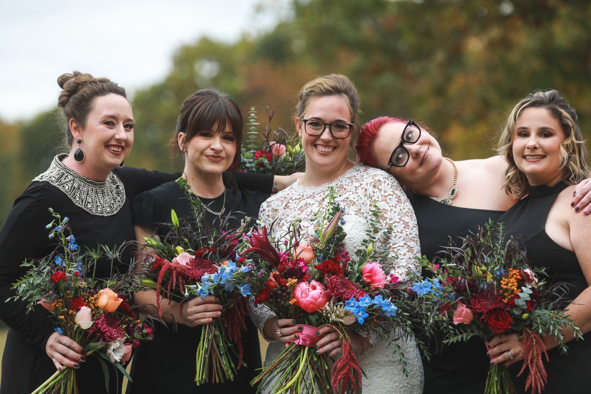 Bridesmaids and bride pose outdoors, holding colorful bouquets. Woman in lace dress smiles, flanked by women in black dresses.