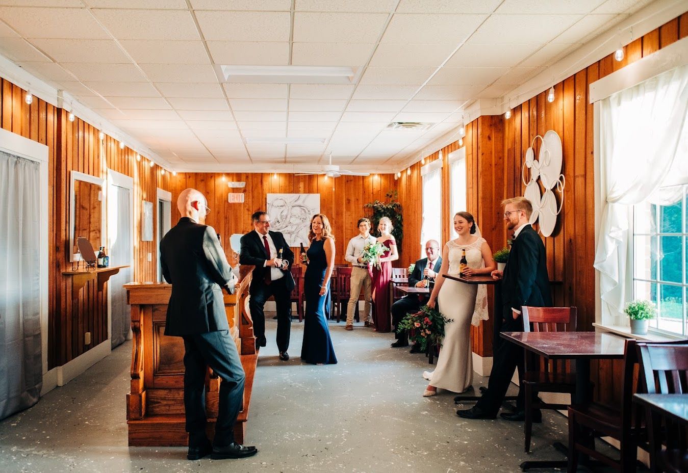 People in formal attire socializing in a wooden-paneled room with windows.