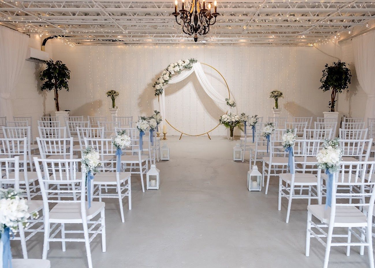 Wedding ceremony setup with white chairs, floral arch, and decorations in a light-filled room.