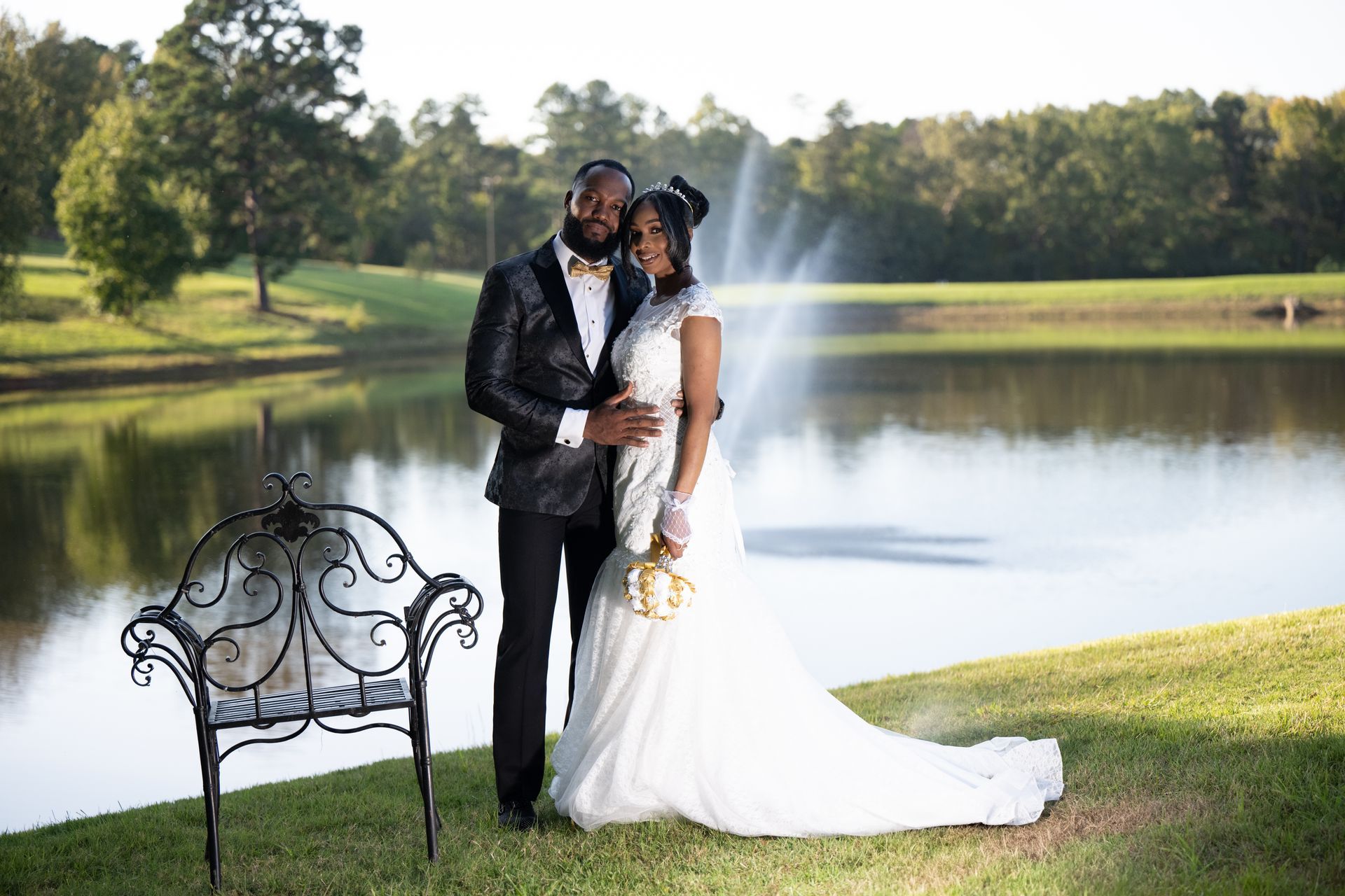 Newlyweds pose by a pond. The bride in white gown, groom in a black suit, standing close together.