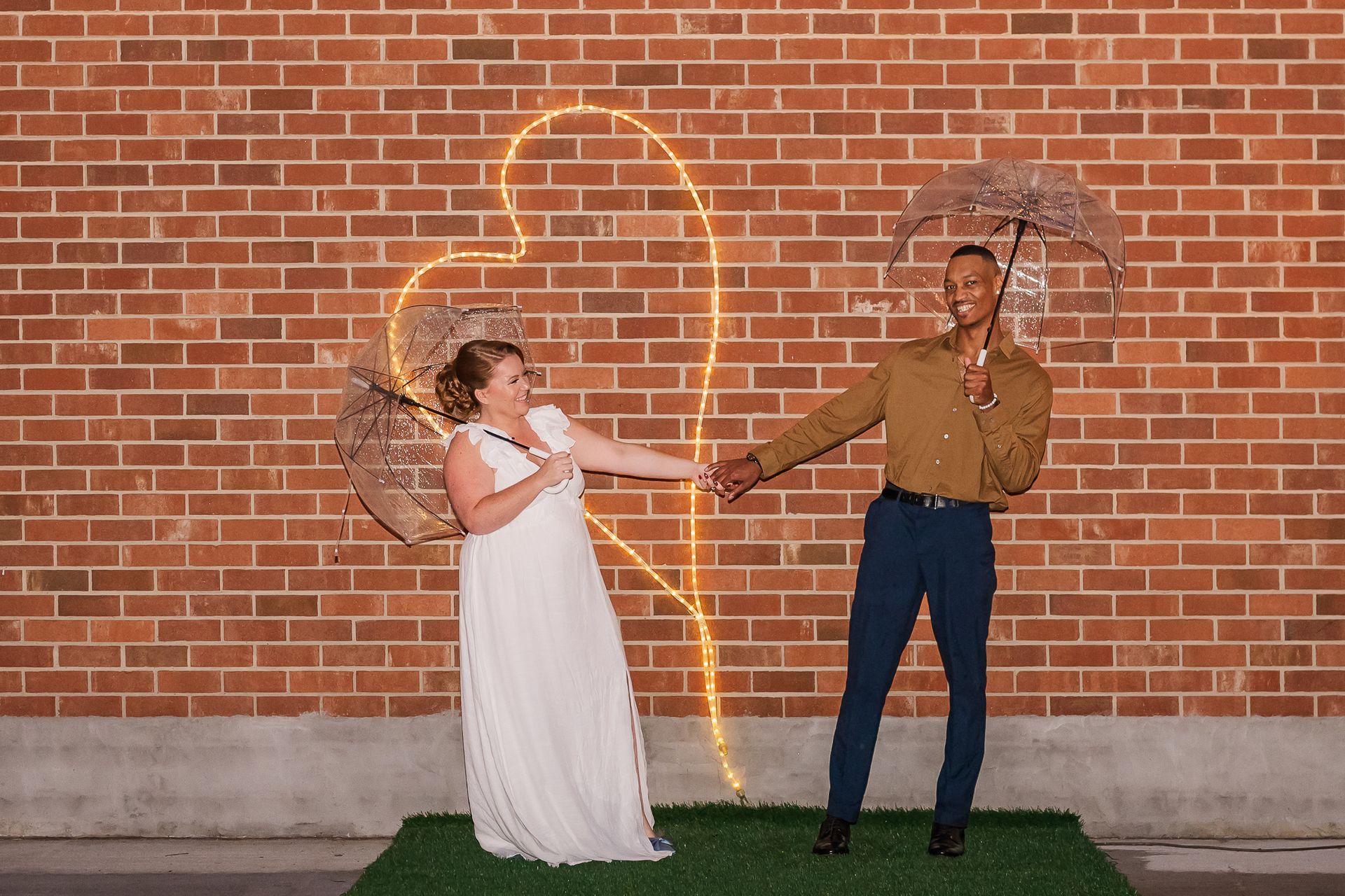 Couple holds hands beneath umbrellas in front of a brick wall with a glowing heart.