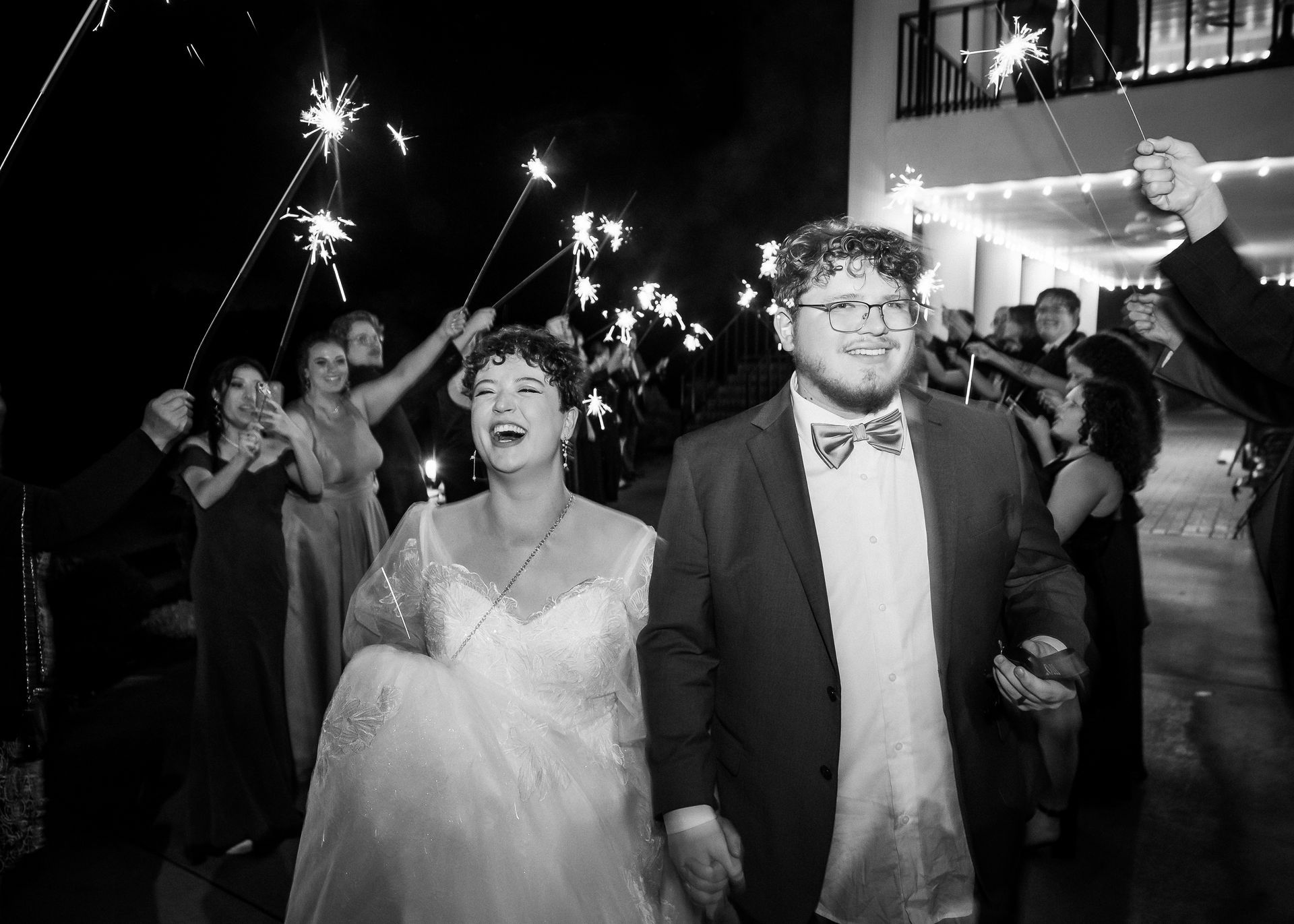 Newlyweds walk through sparkler-lit exit, smiling. Guests with sparklers line a walkway at night.