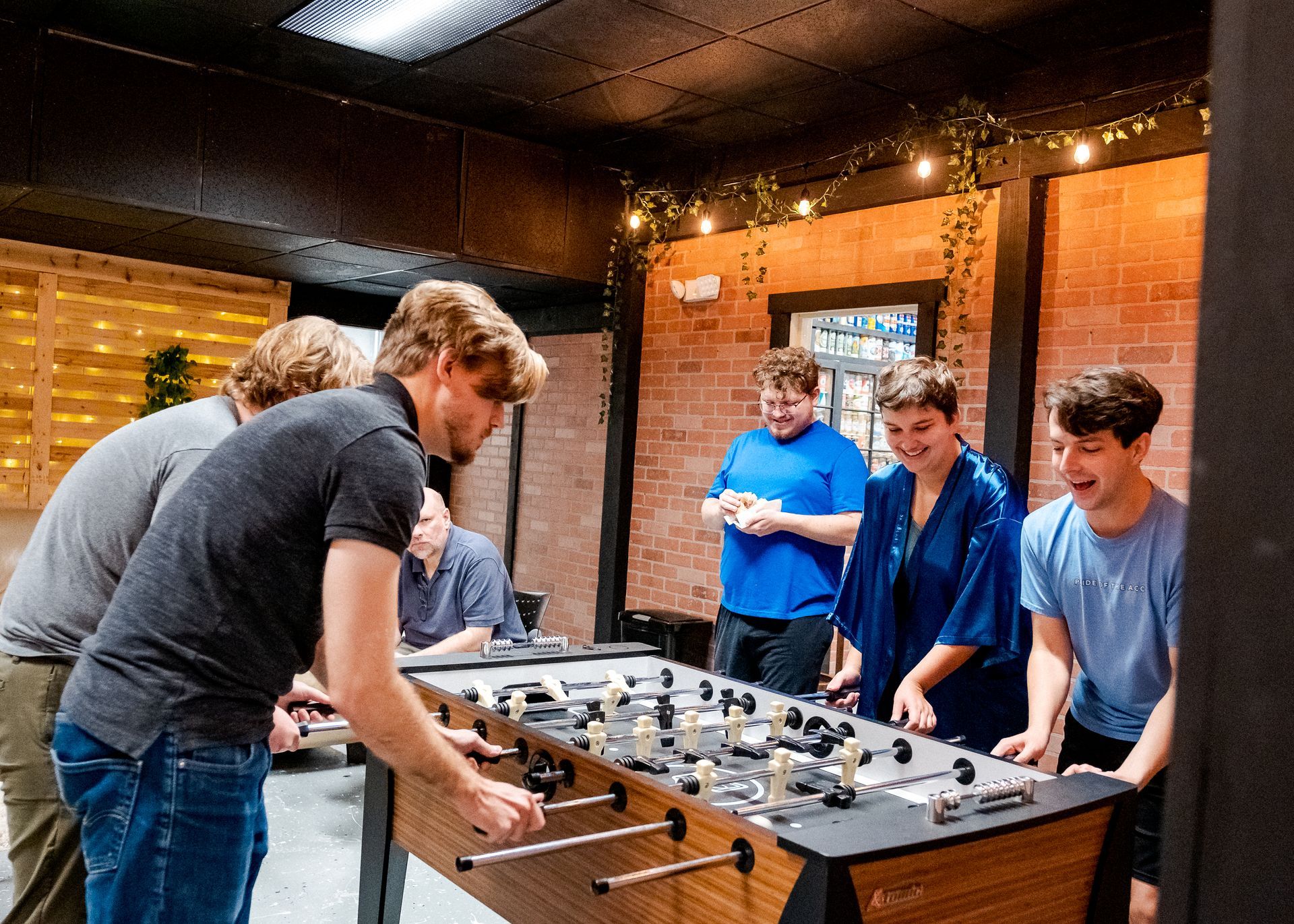 People playing foosball in a room; some watching and smiling. Brick and wood walls, string lights.