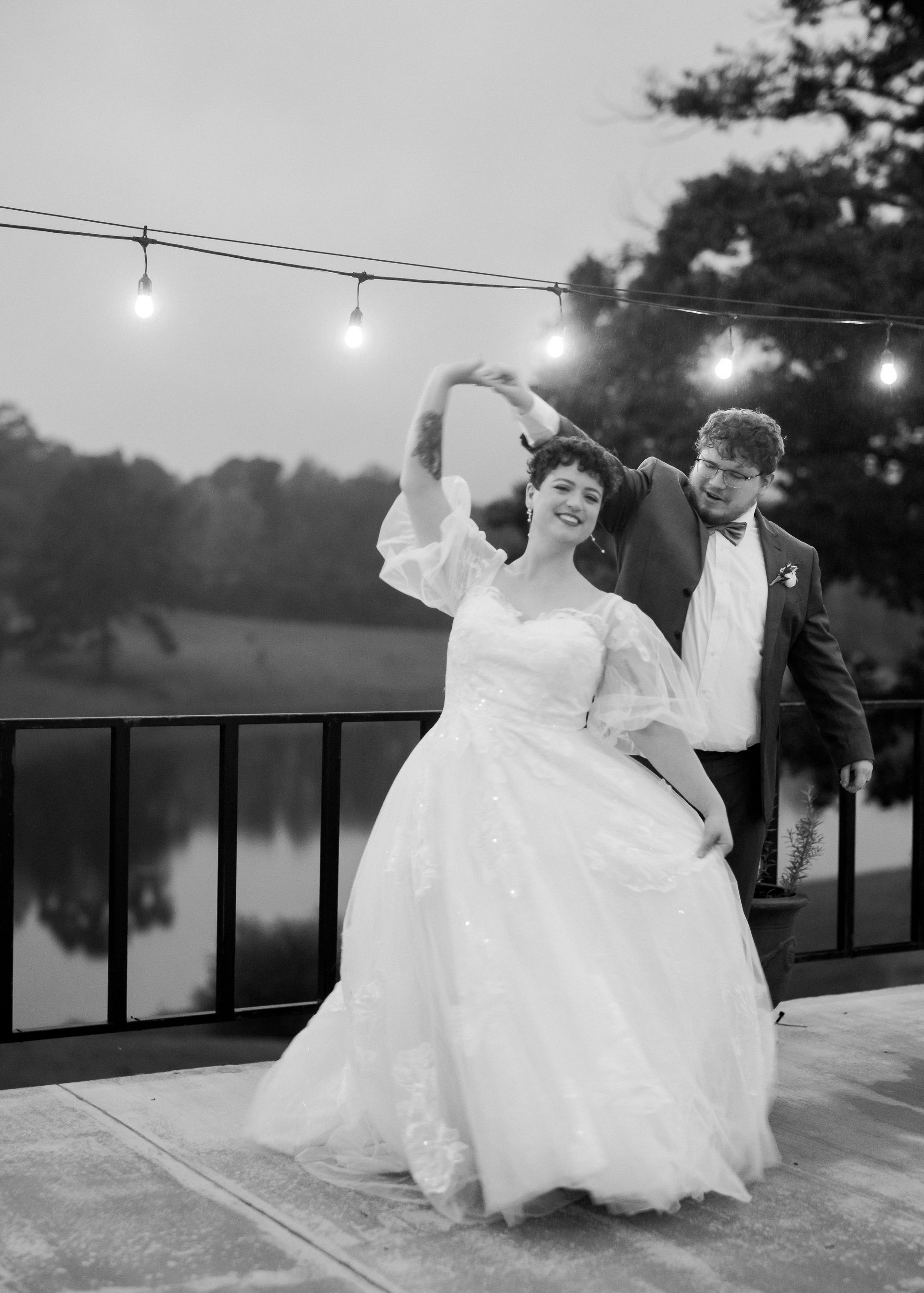 Couple dancing on a deck, bride in a wedding gown, lights strung overhead, lake in background.
