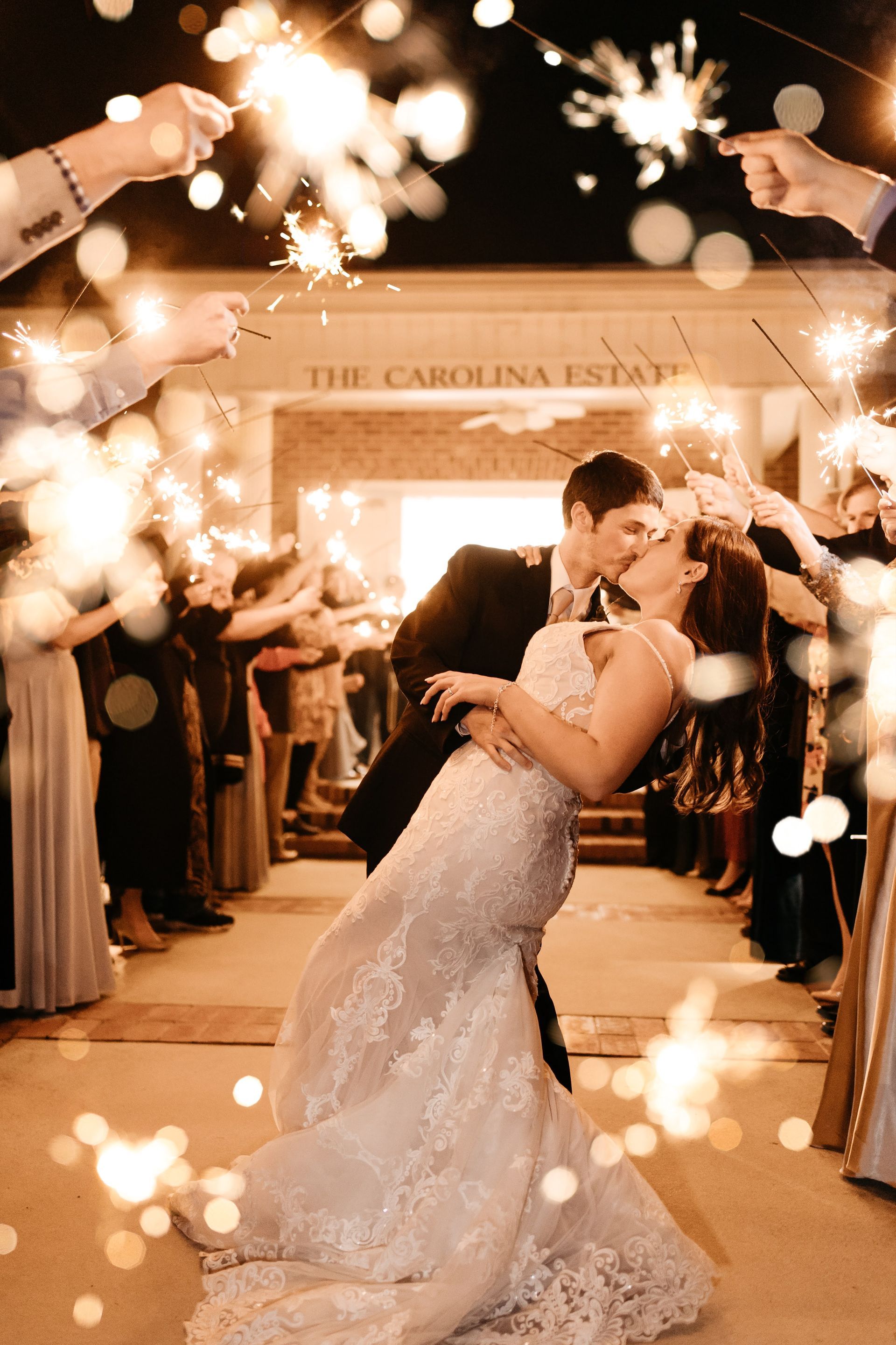 Newlyweds kiss as guests hold sparklers at their wedding. The Carolina Estate building is in the background.