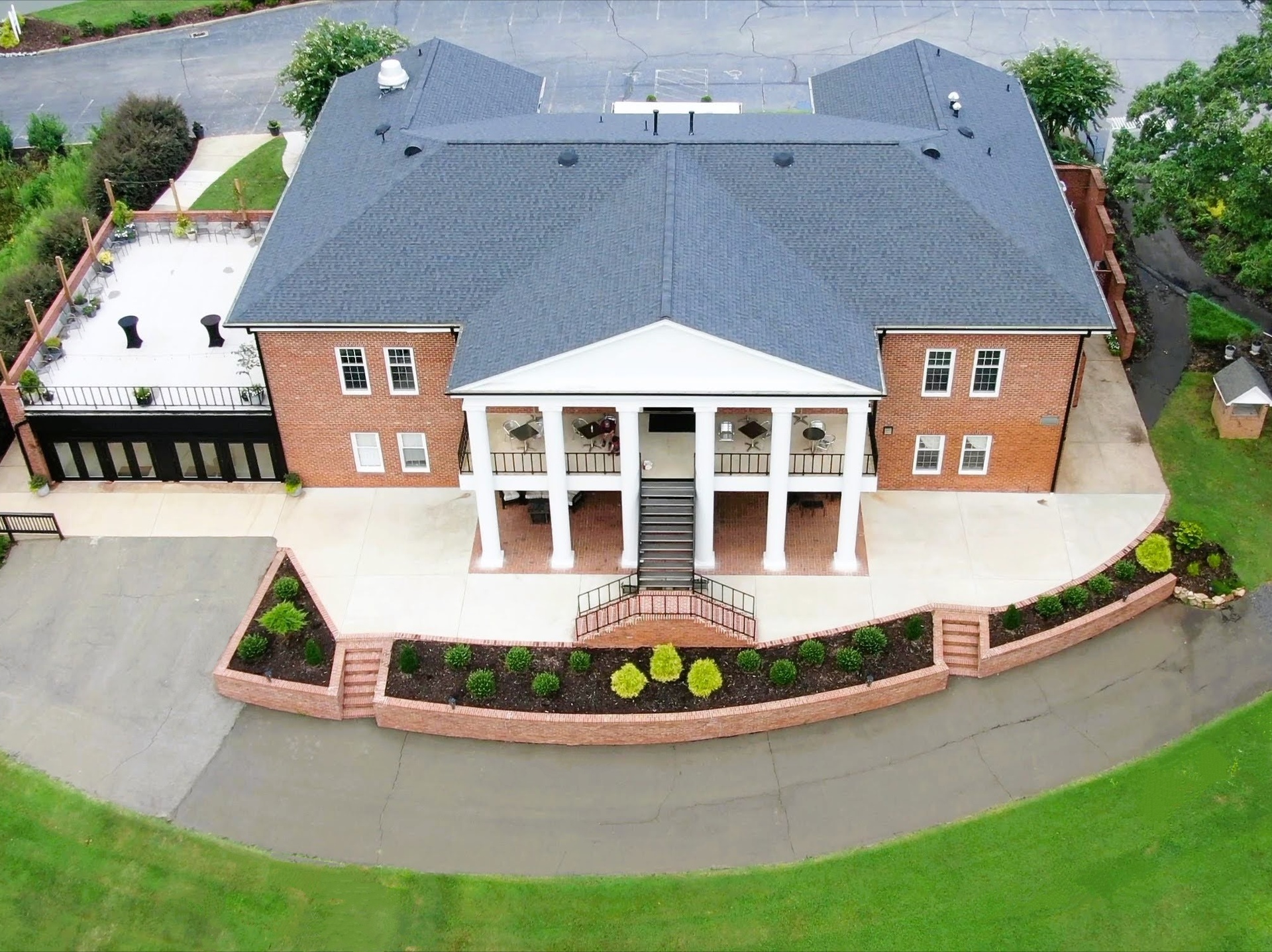 Aerial view of a brick building with a columned entrance, a dark gray roof, and a surrounding paved area.