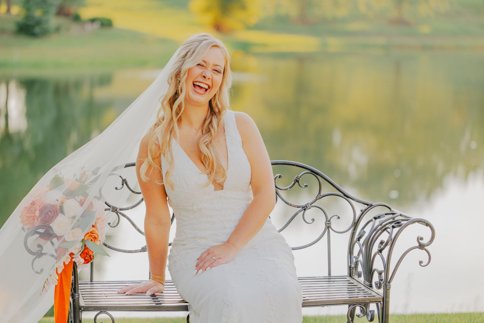 Bride laughs on a bench by a lake, wearing a white dress and veil, holding a bouquet.