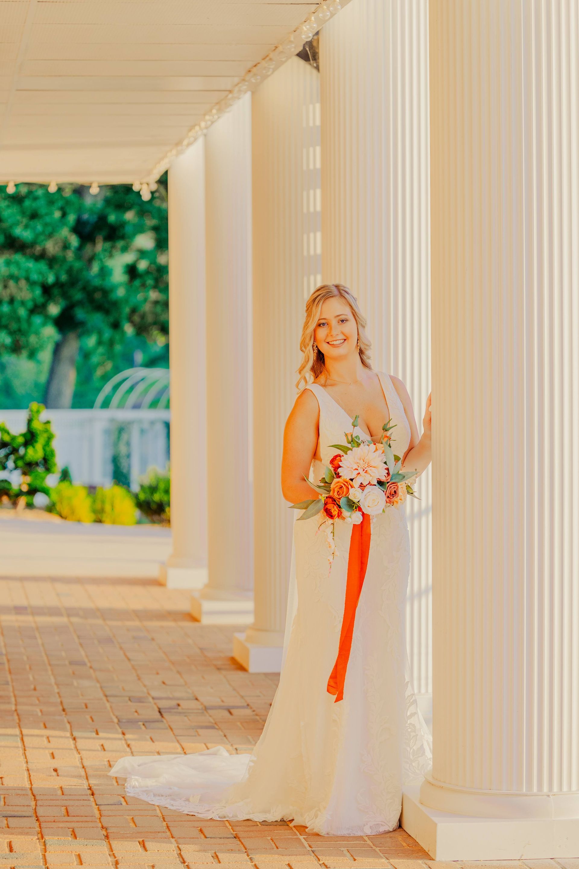 Bride in a white gown holding a bouquet, standing by a white pillar. Outdoor setting with sunlight.