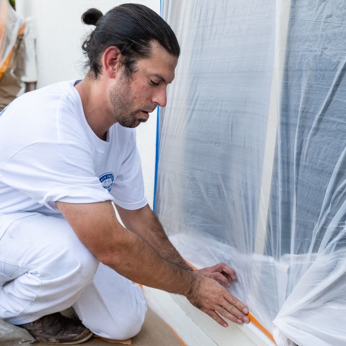 A man is kneeling down and covering a window with plastic