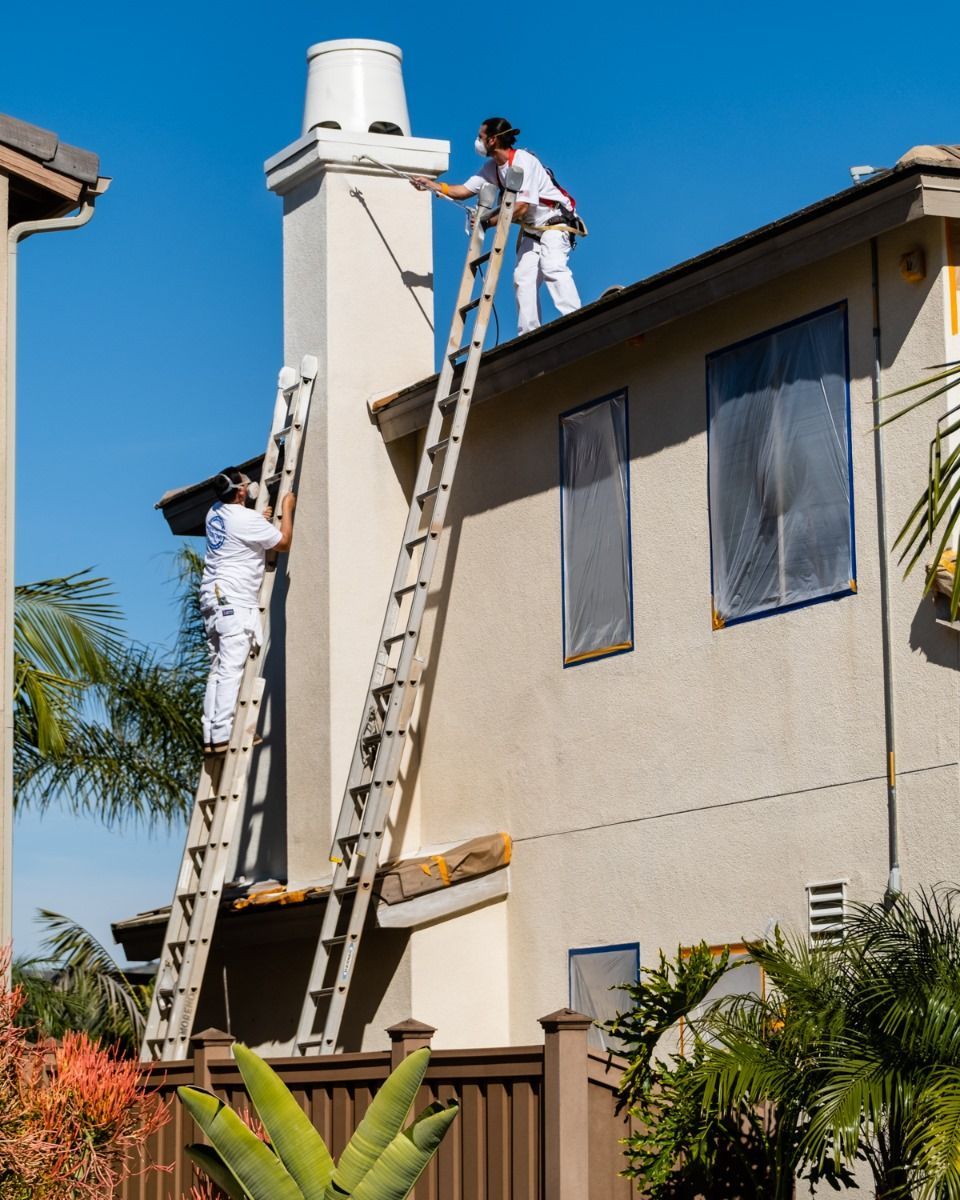 A man on a ladder is painting a chimney on the side of a house.