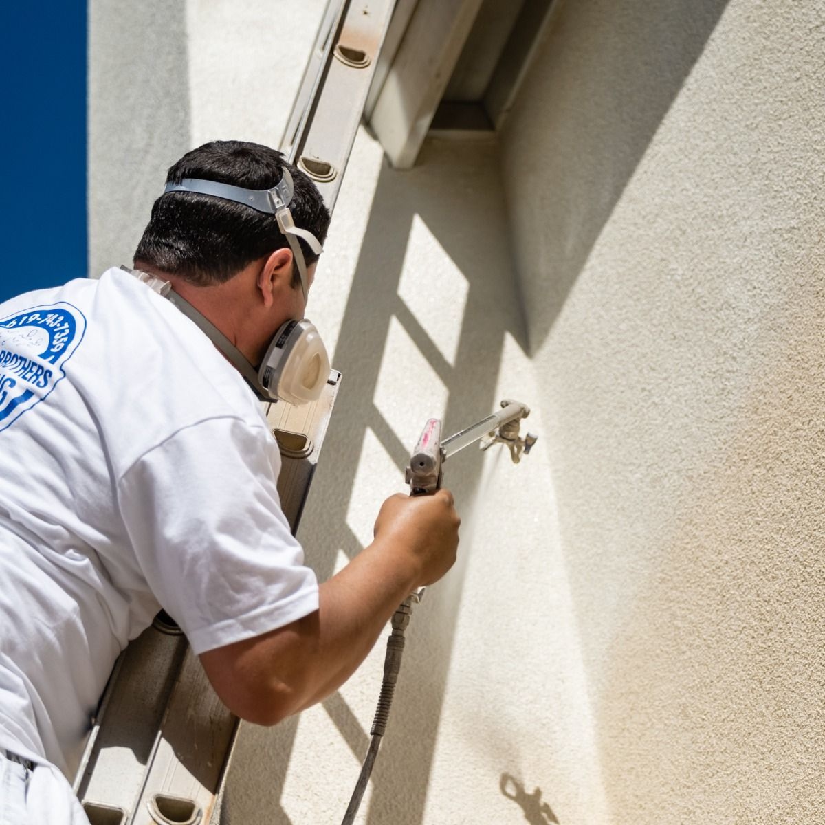 A man wearing a white shirt with a blue logo on the back is painting a wall