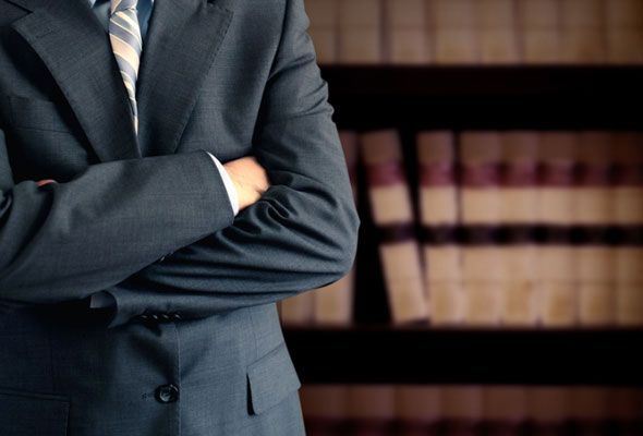 A man in a suit and tie is standing with his arms crossed in front of a bookshelf