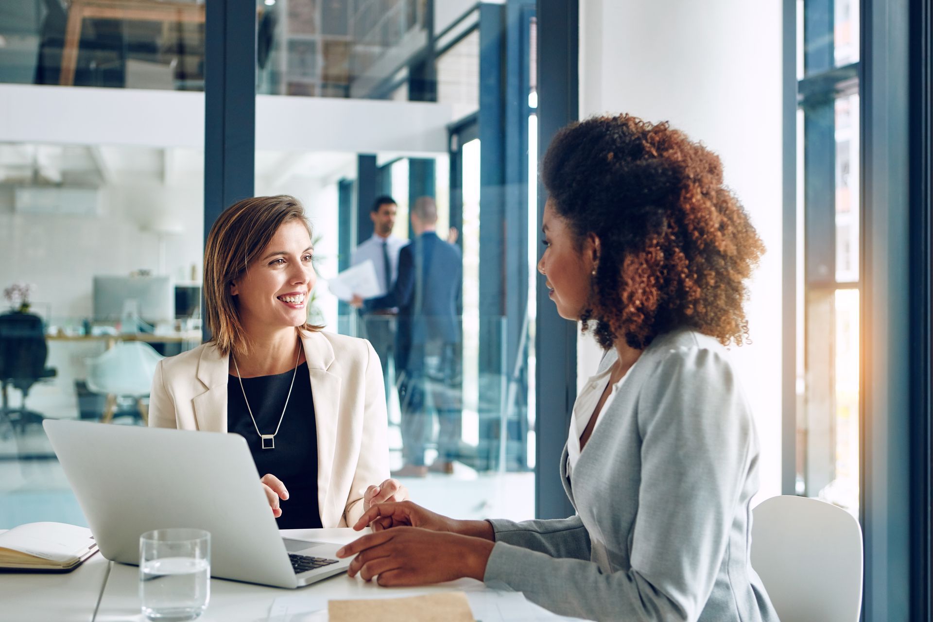 Two coworkers talking at a laptop in a bright glass-walled office.