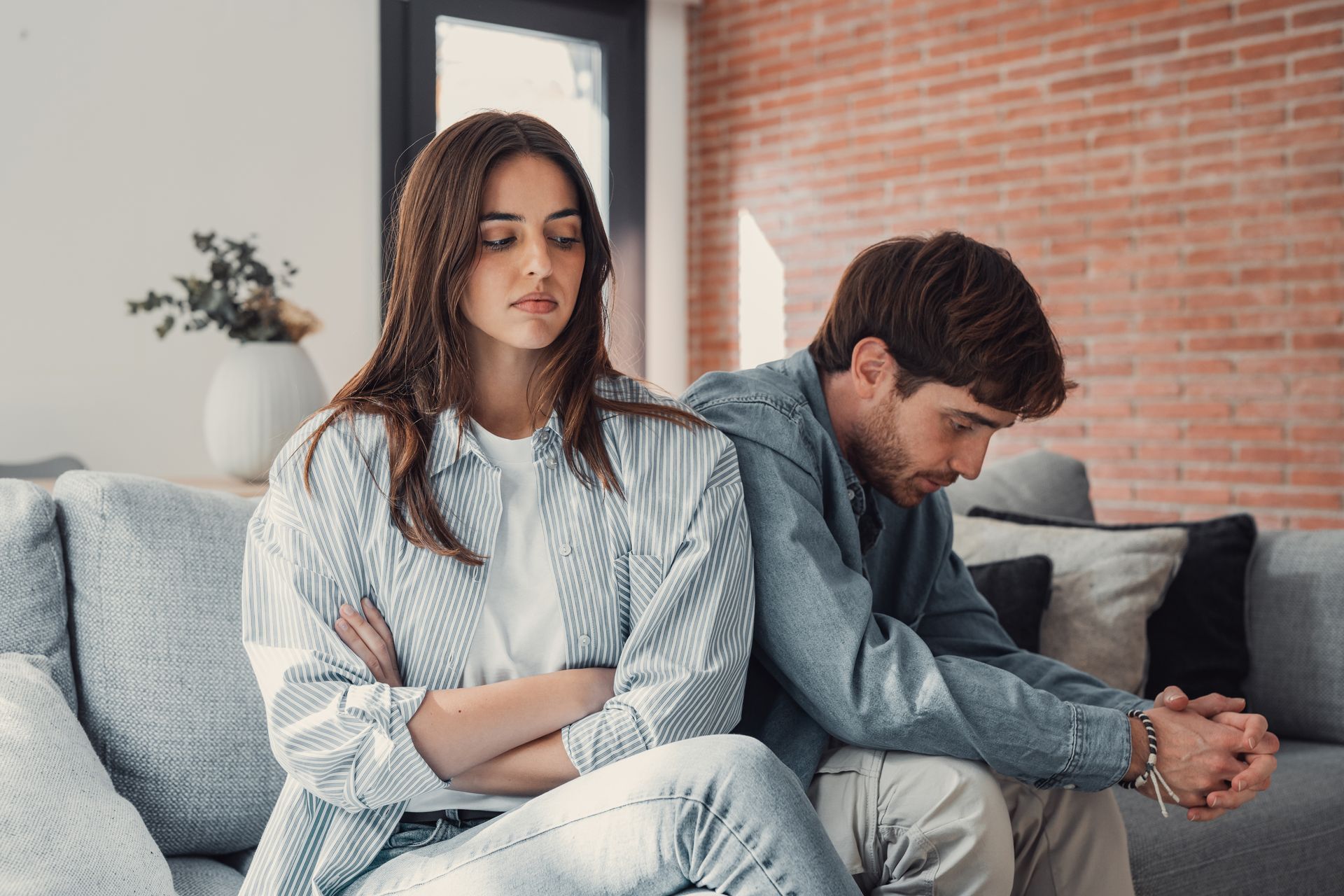 Two people sit apart on a couch, looking down with crossed arms in a tense living room.