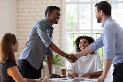 Two people shake hands across a conference table while others smile in a bright meeting room.