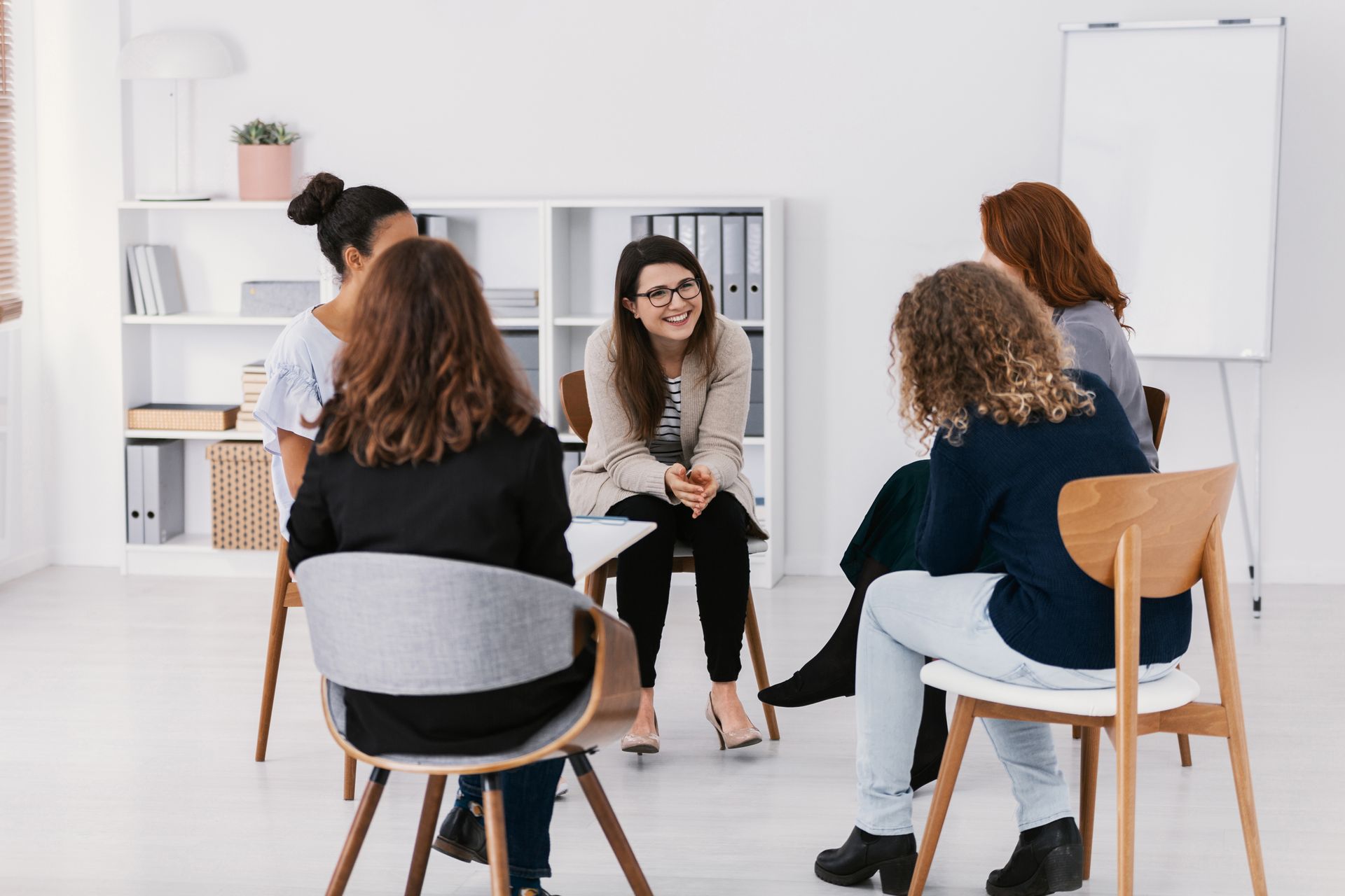 Small group discussion in a bright meeting room, with people seated in a circle and a presenter at a whiteboard.