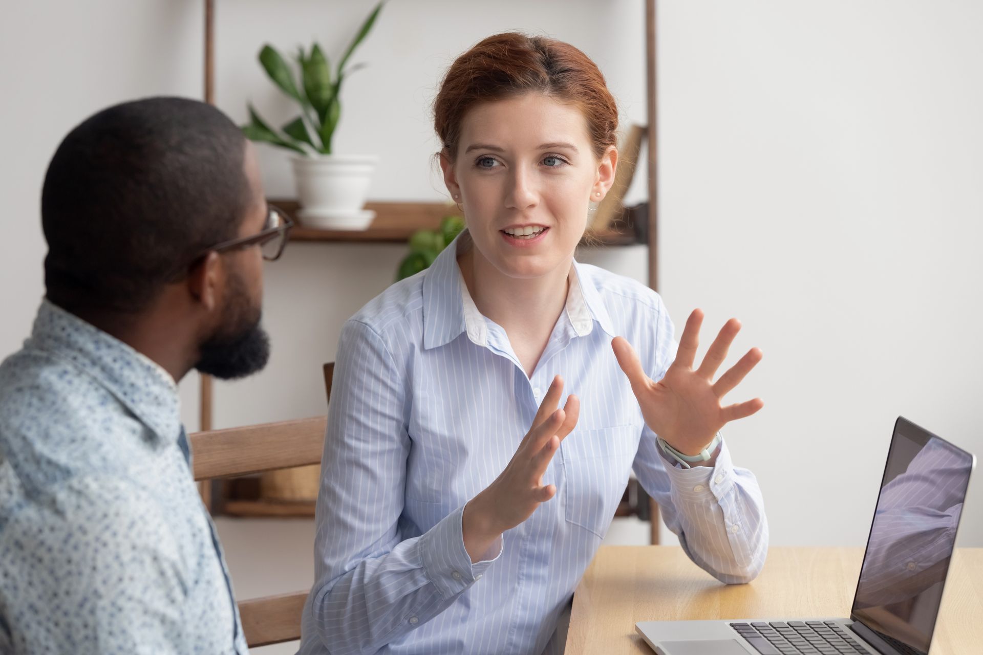 Two coworkers talking at a desk, with one gesturing beside a laptop in a bright office