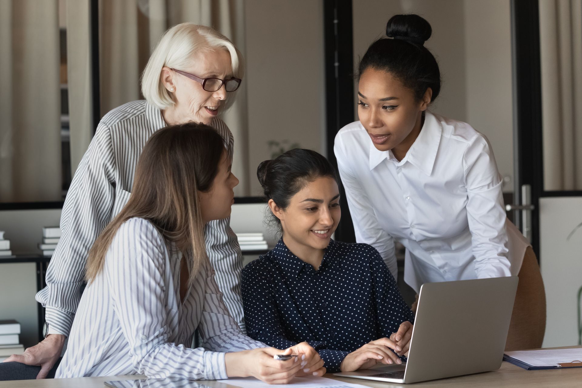 Four coworkers gathered around a laptop in a bright office, discussing something together.