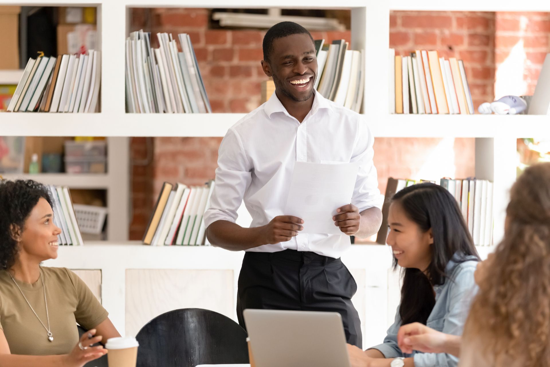 Colleagues chatting around a laptop in a bright office with bookshelves, one man standing and smiling.