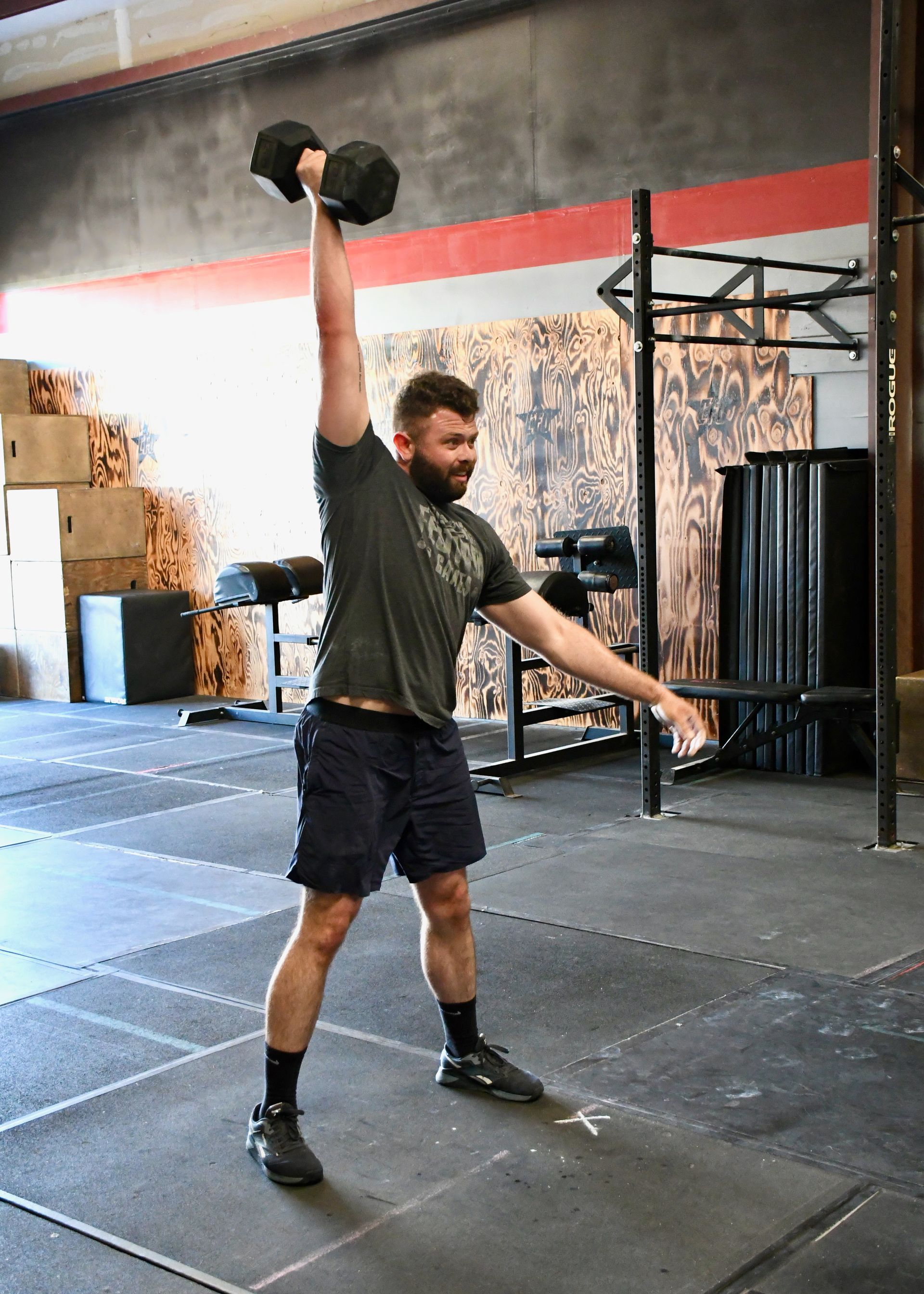 Woman in orange tank top lifts a dumbbell overhead in a gym. A second woman watches.