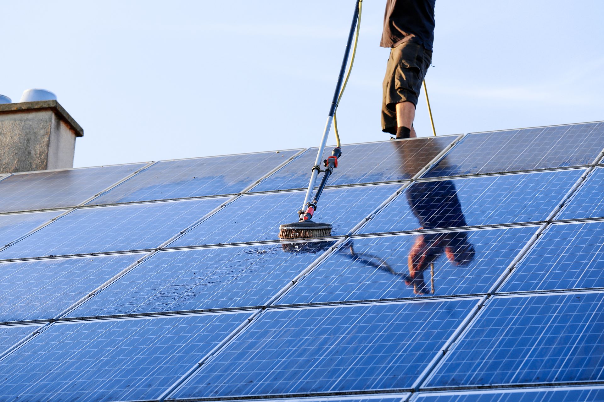 Worker cleaning blue solar panels on a sloped rooftop with a hose